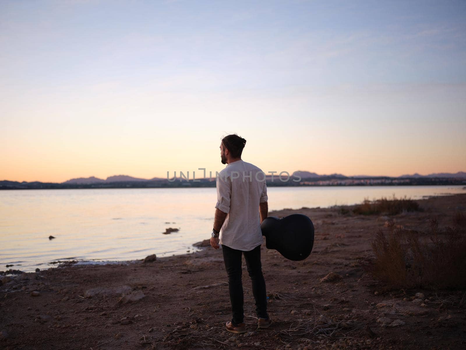 man gazing at the horizon with a guitar in front of the pink lake at sunset, horizontal landscape