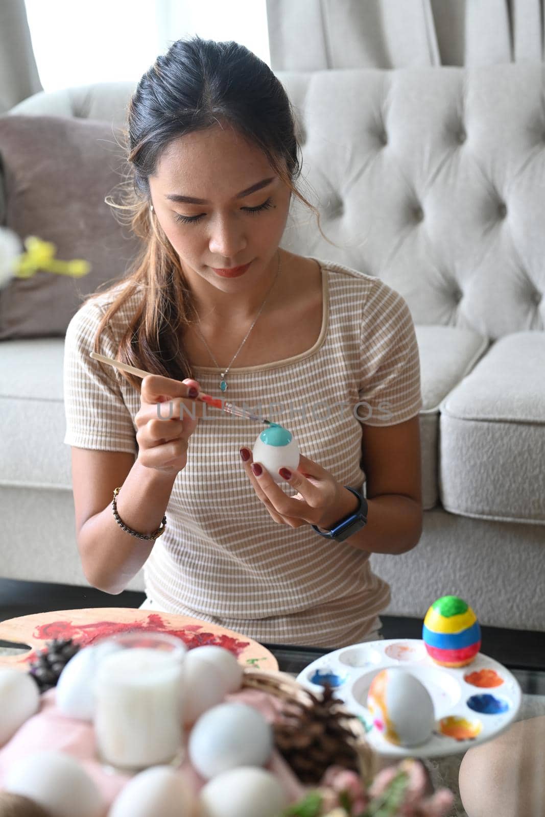 Attractive young woman sitting in living room and painting egg for Easter festivity.