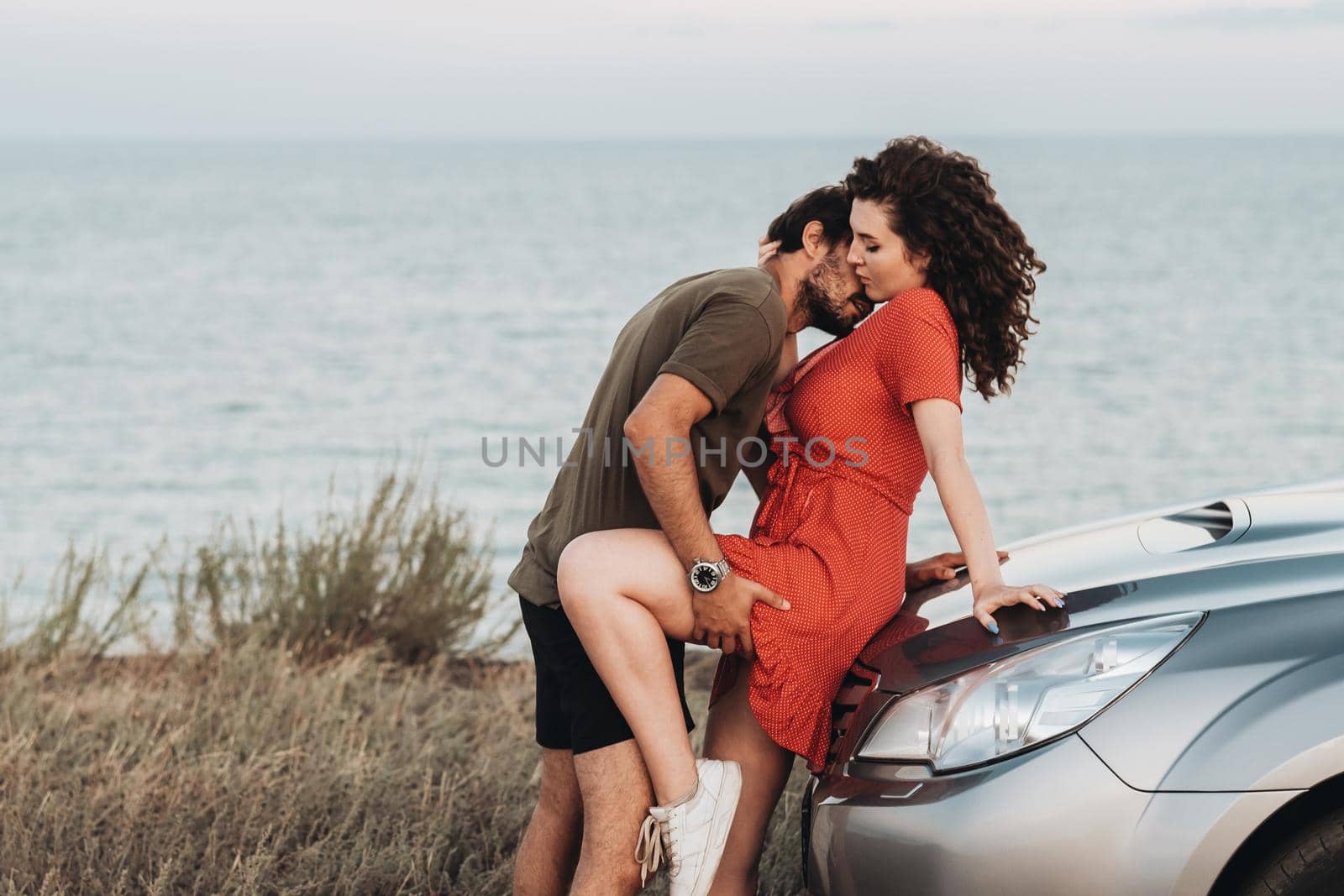 Young Couple on Road Trip, Man Intimate Kissing Woman in the Neck on the Hood of Their SUV Car on Background of Sea at Sunset