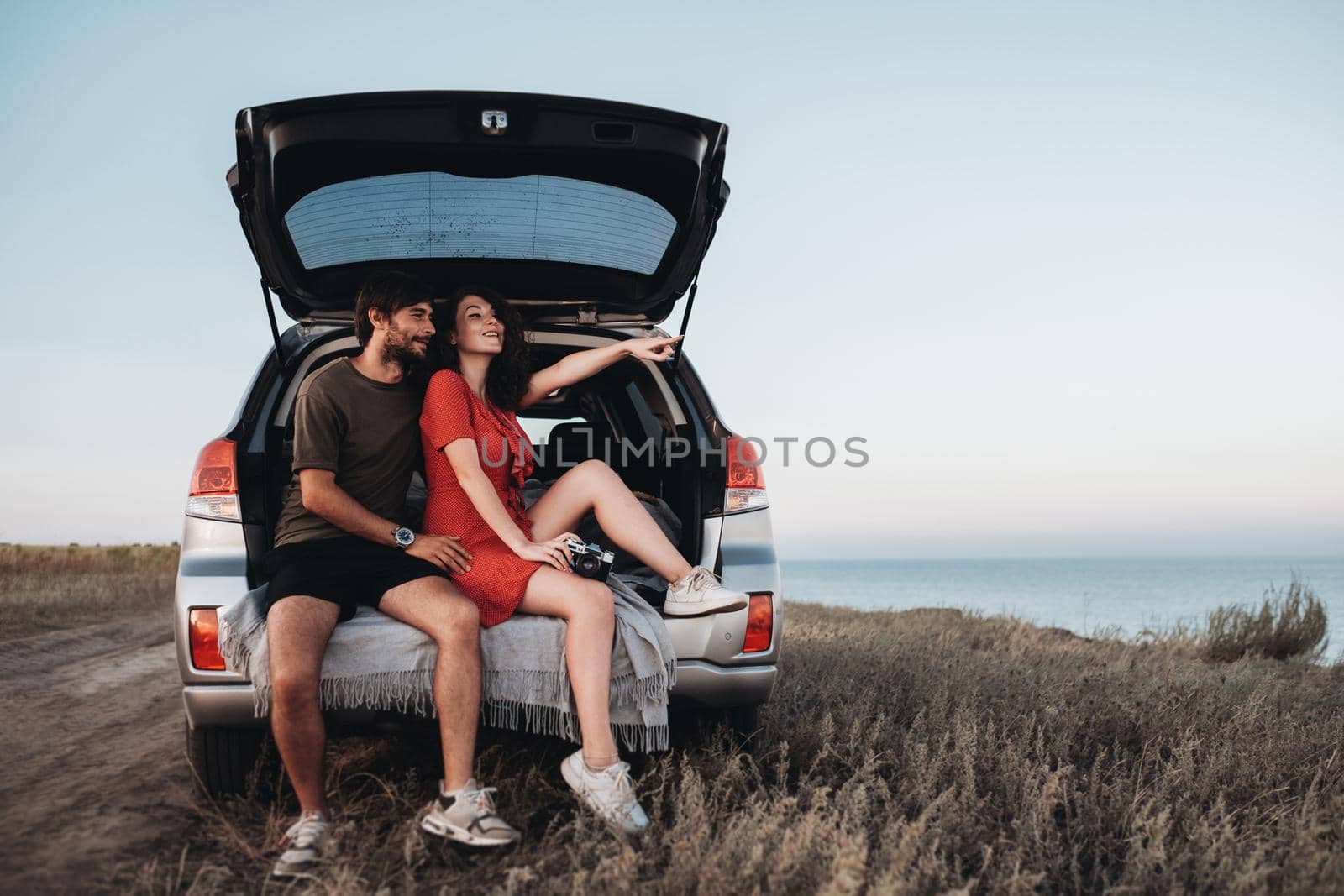 Man and Woman Sitting Inside Opened Trunk of Their SUV Car, Young Couple Enjoying Road Trip Along Sea at Sunset by Romvy