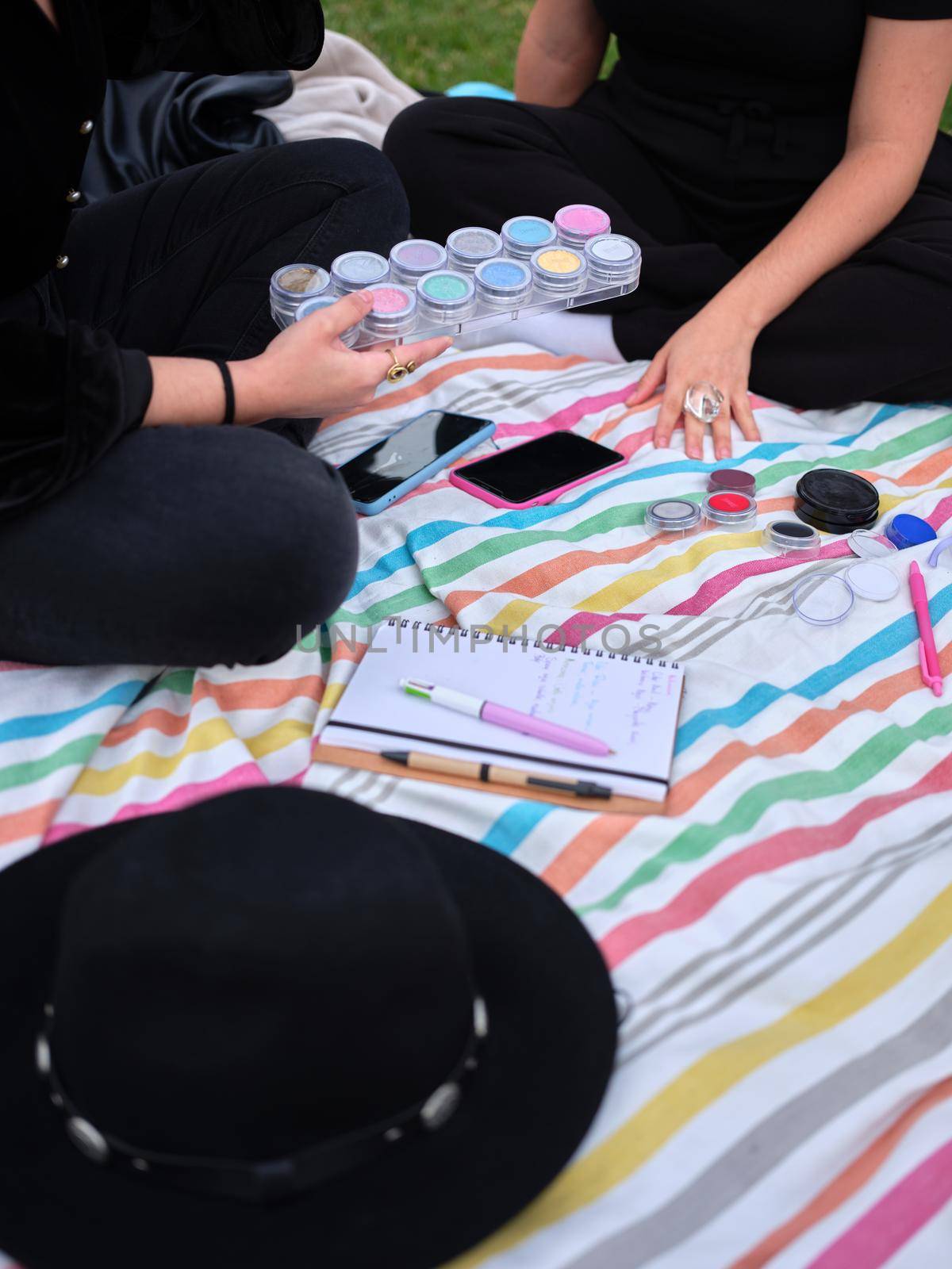 two young women sitting with legs crossed, one of them picking up the paints to choose which one to paint with