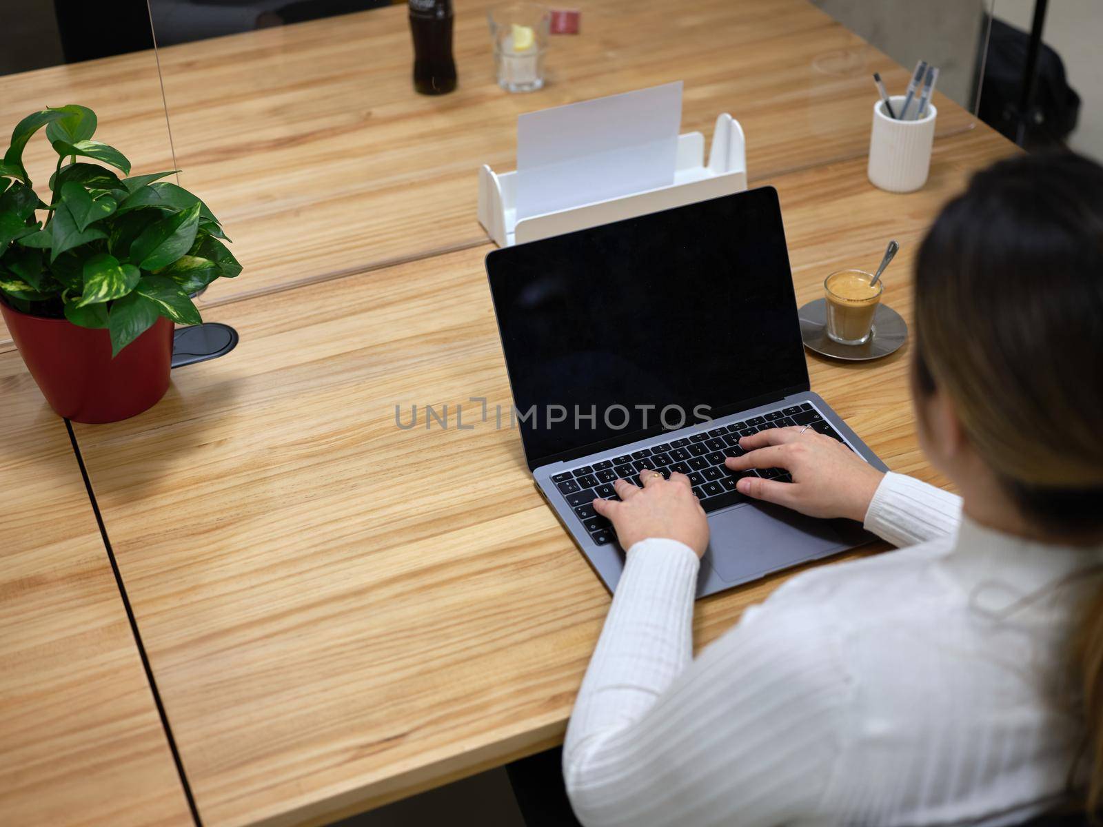 view from behind of a woman using her laptop in a coworking space, plant and a coffee over the desk