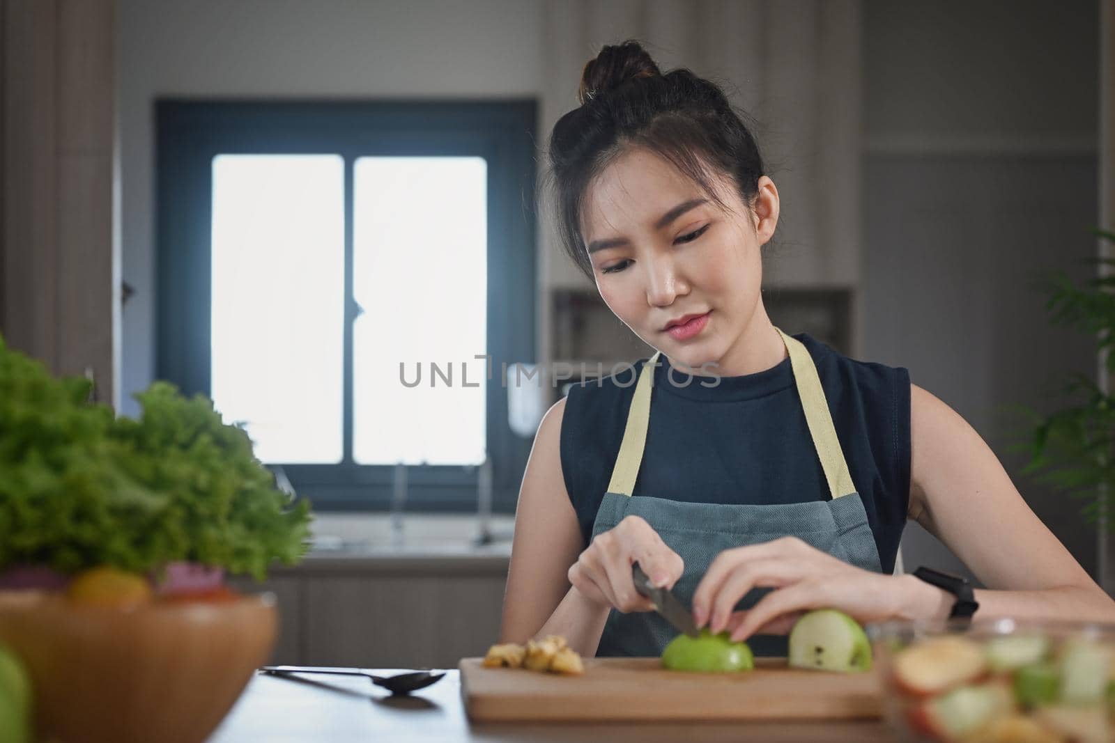 Asian woman in apron sitting at kitchen table and cutting apple on wooden cutting board during preparing healthy salad.