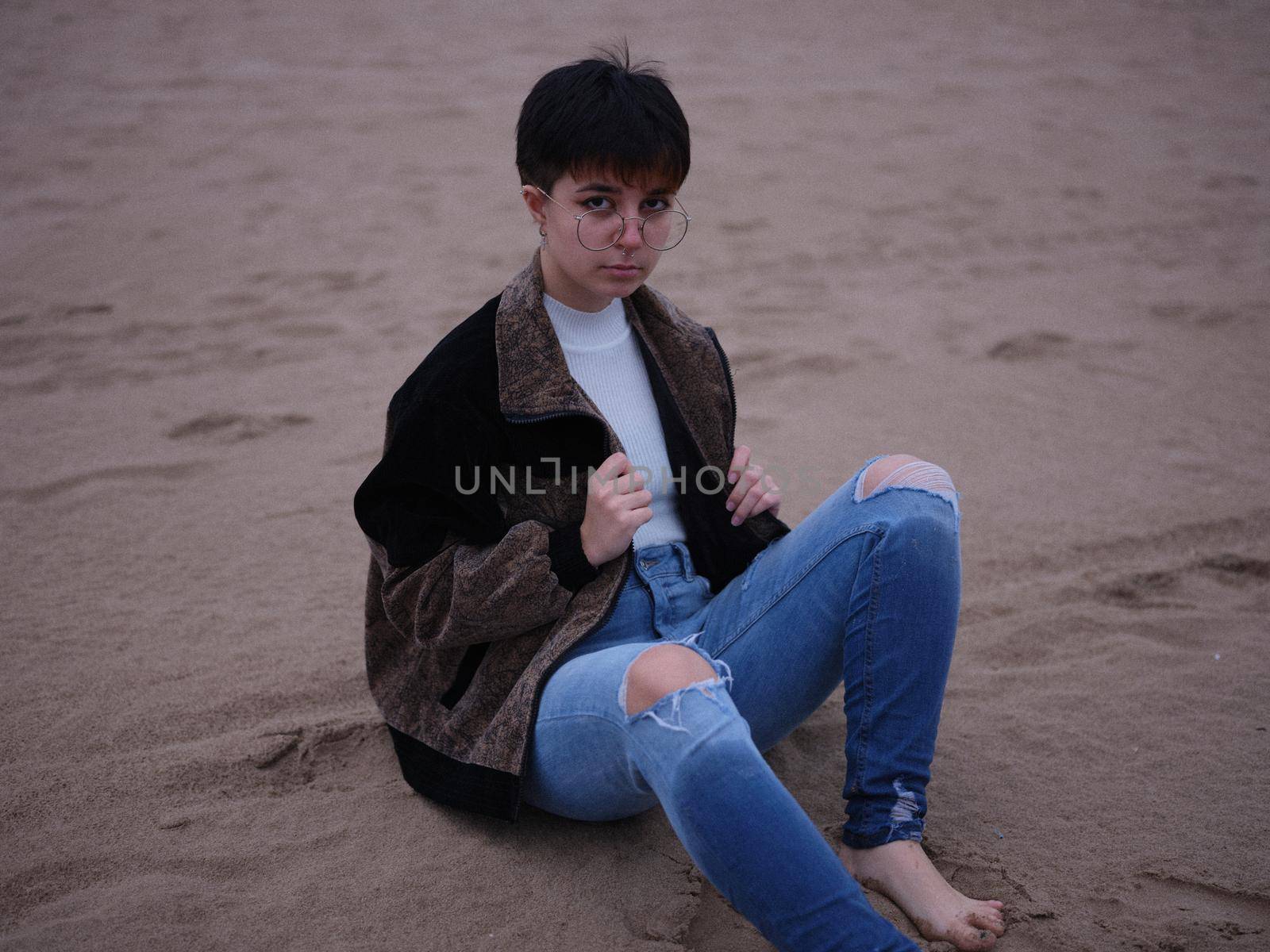 transgender person sitting on the sand with a jacket and glasses, horizontal portrait