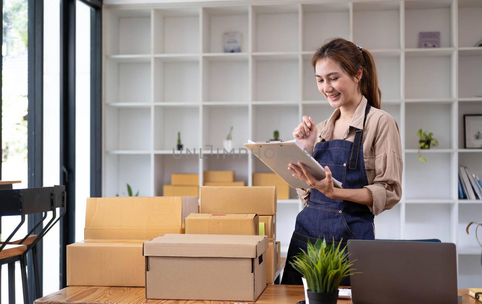 Beautiful smart Asian young entrepreneur business woman owner of SME checking product on stock and write on clipboard working at home. Small business owner at home office concept..