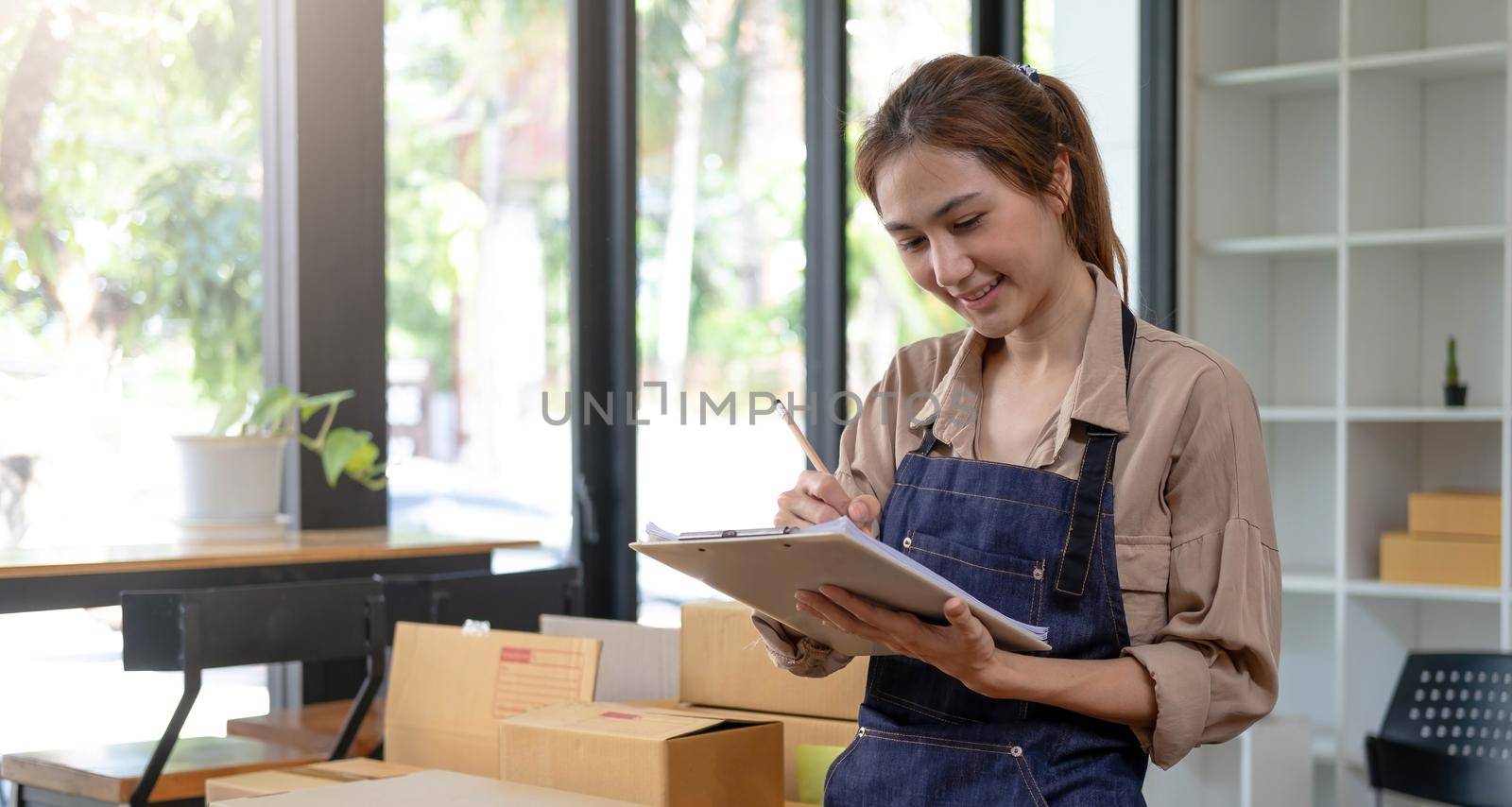 Beautiful smart Asian young entrepreneur business woman owner of SME checking product on stock and write on clipboard working at home. Small business owner at home office concept..