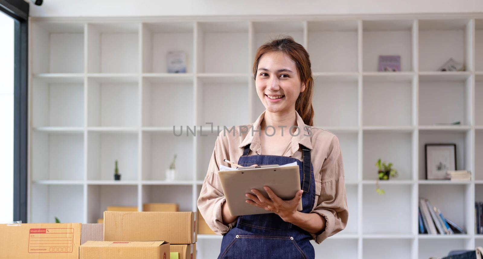 Beautiful smart Asian young entrepreneur business woman owner of SME checking product on stock and write on clipboard working at home. Small business owner at home office concept. by wichayada