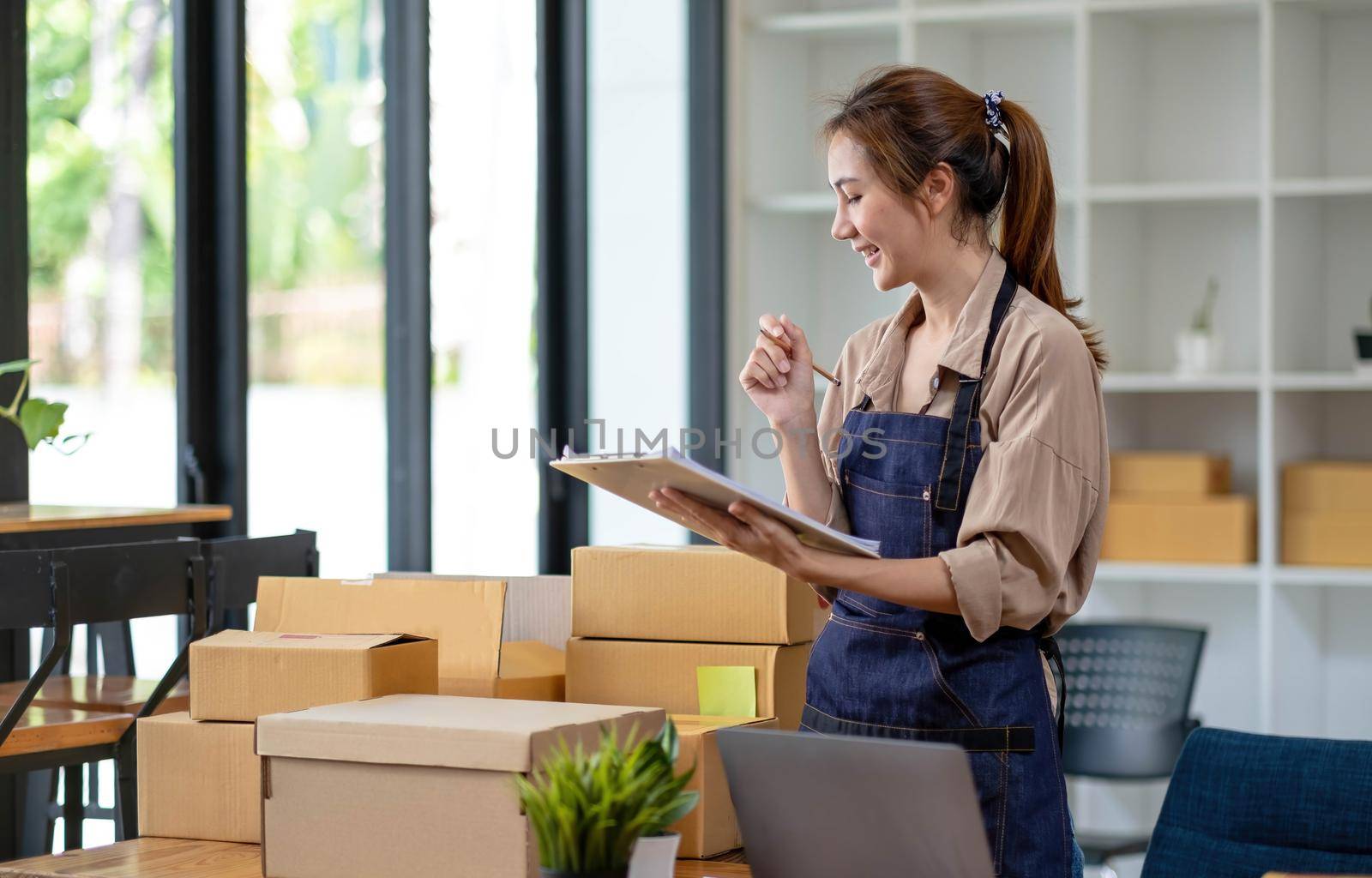 Beautiful smart Asian young entrepreneur business woman owner of SME checking product on stock and write on clipboard working at home. Small business owner at home office concept..