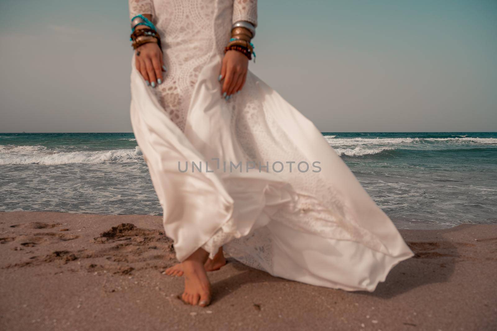 Model in boho style in a white long dress and silver jewelry on the beach. Her hair is braided, and there are many bracelets on her arms