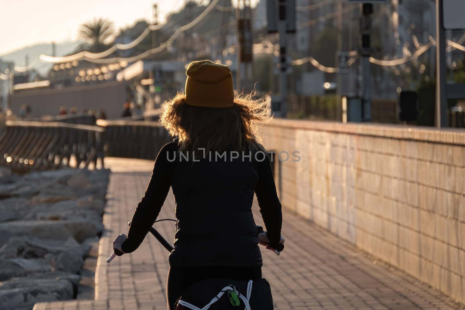 Young girl in a yellow hat with curly hair fluttering in the wind riding a bicycle at sunset on the promenade road of Maresme, Catalonia, Spain.