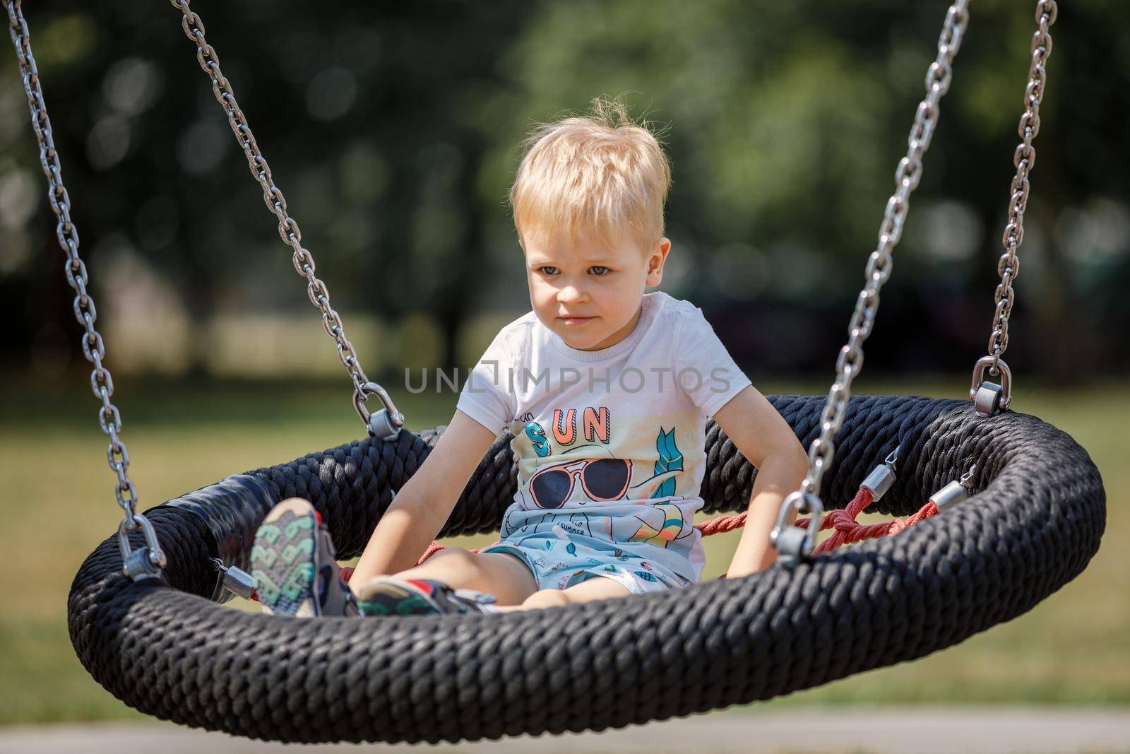 Little blonde boy playing happily on large nest swing at city playground. by Lincikas