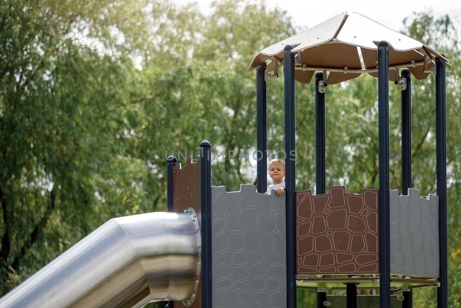 A little boy in the high observation tower of the playground prepares to slide through a metal tunnel. Beautiful background of green, tall trees.