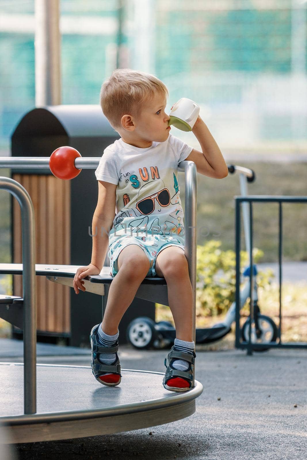 Little boy rides carousel in playground outside. Cute little child. Concept of kindergarten and childhood. Metal carousel for children in summer in the park. Vertical.