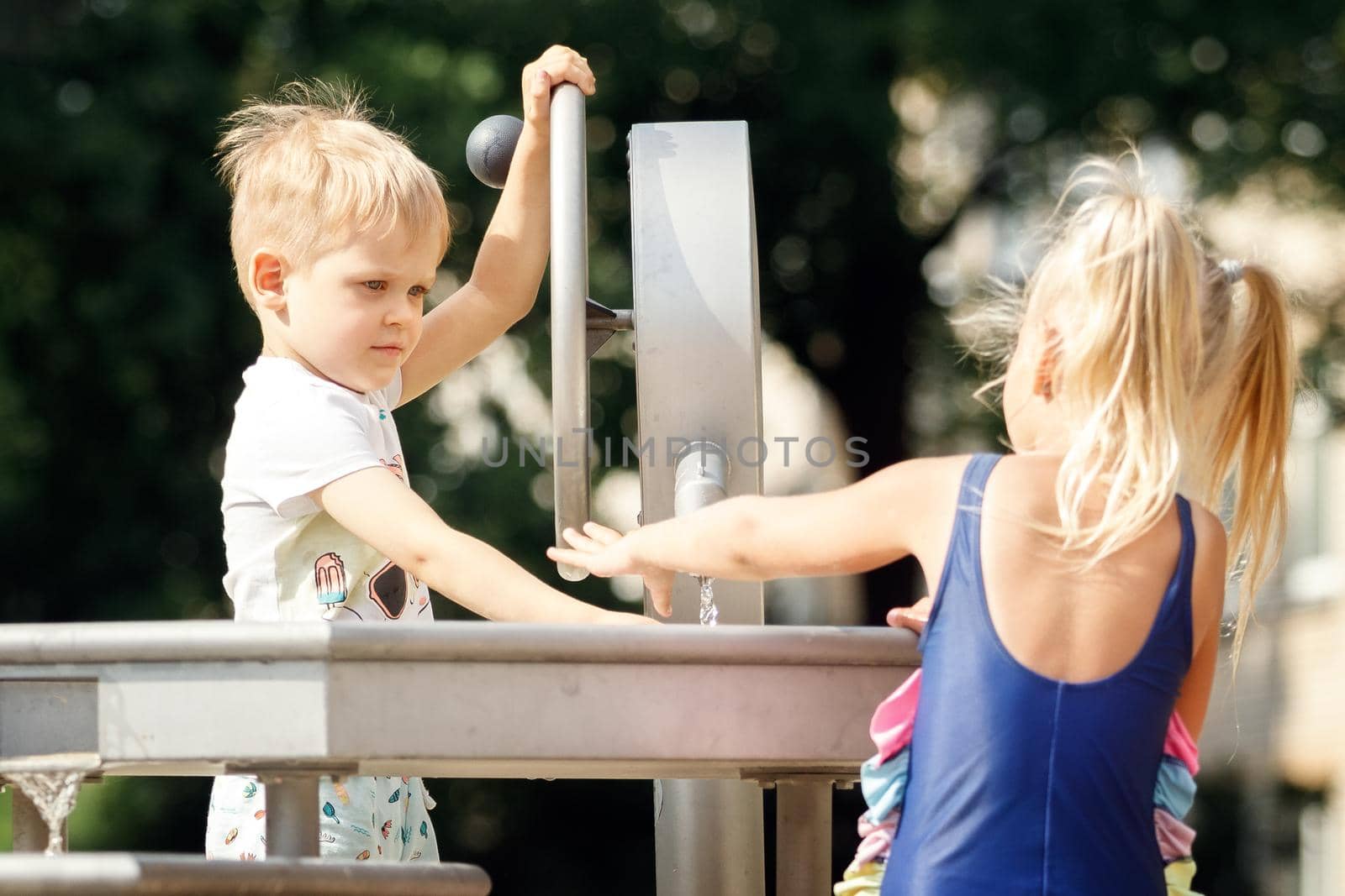 A happy boy and a cute girl in a blue bathing suit play with a water tap in a city park. Horizontal.