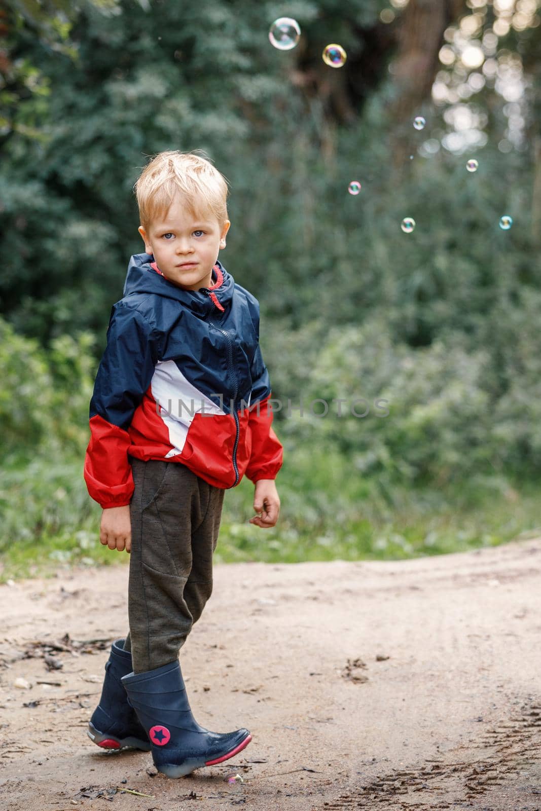 Portrait of a blonde cute boy posing in the nature of autumn. The child wears a colorful waterproof jacket and rubber boots.