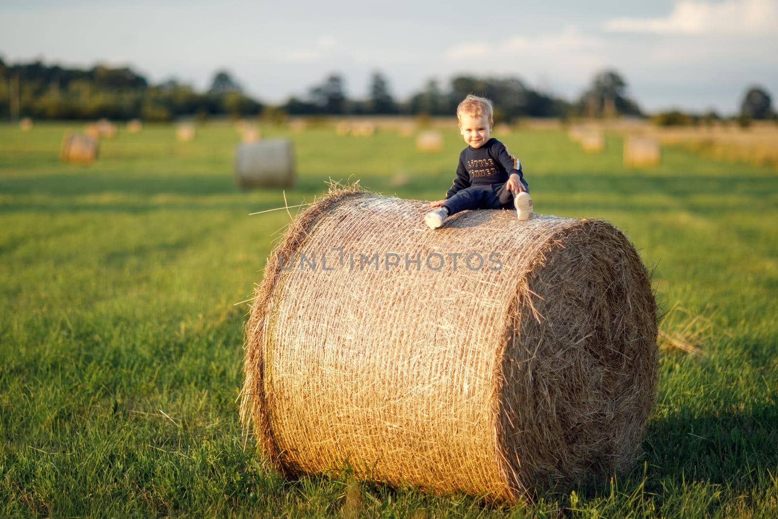 The little boy sits on a large roll of hay in a green meadow, posing beautifully with a smile.