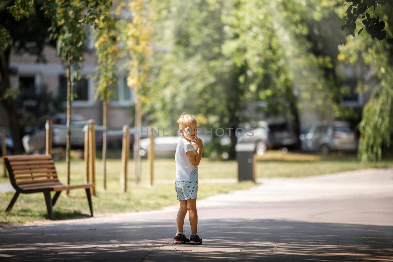 A little boy is standing in the shade of trees in a city park, the child is alone he is afraid of getting lost.