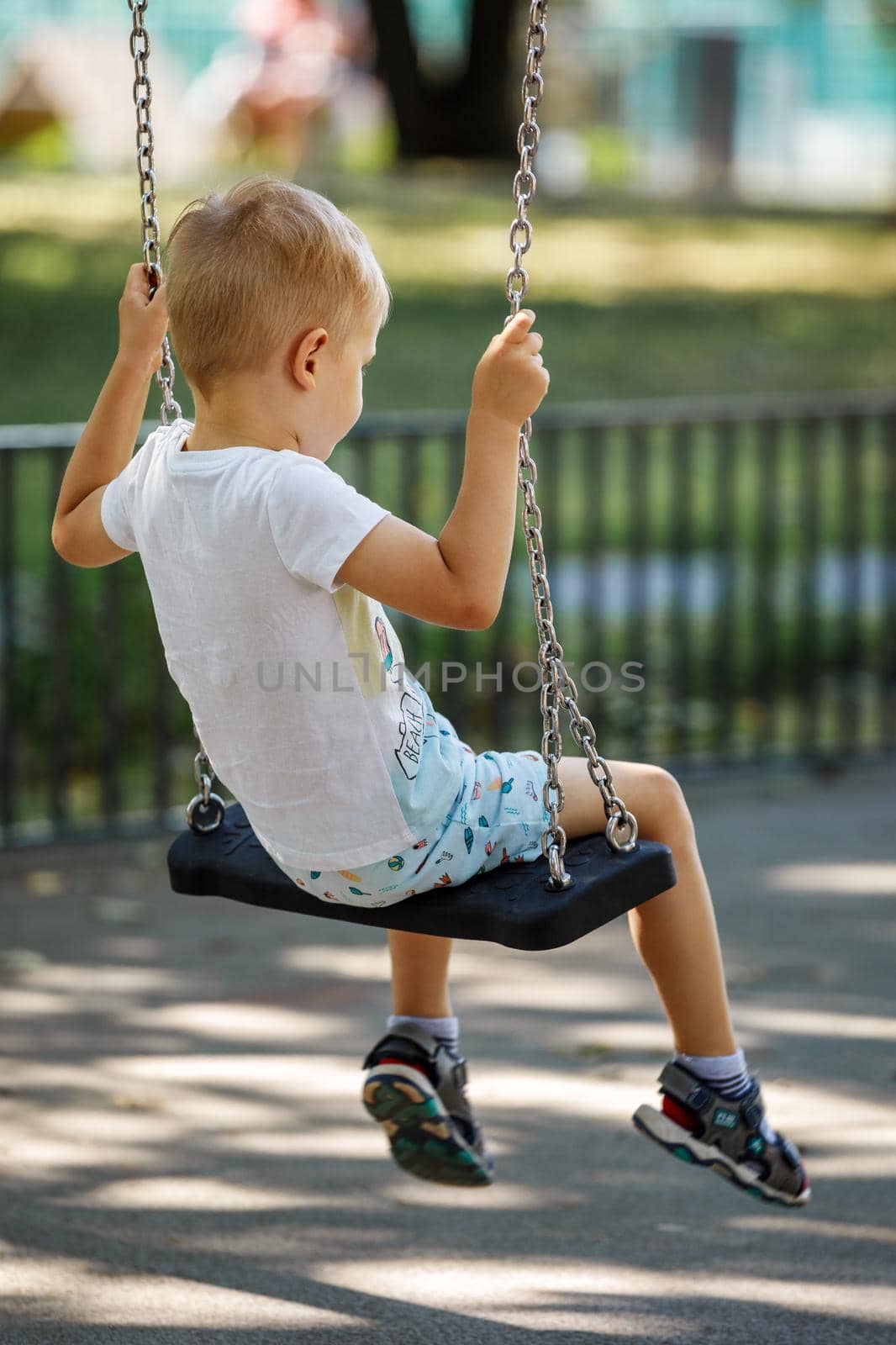 A blond boy in a white T-shirt swings on a swing in the park on a summer day, photo from behind by Lincikas
