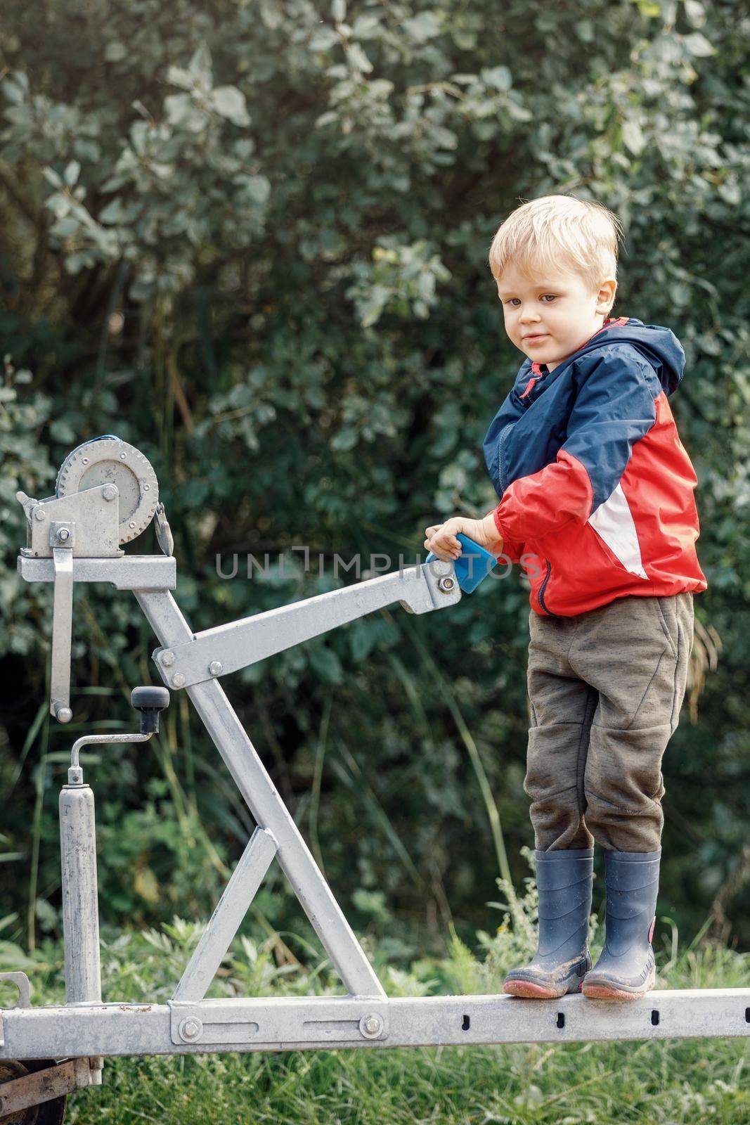 The little happy boy with rubber boots climbs on boat trailer winch and poses for the camera against a background of green foliage. Vertical photo.