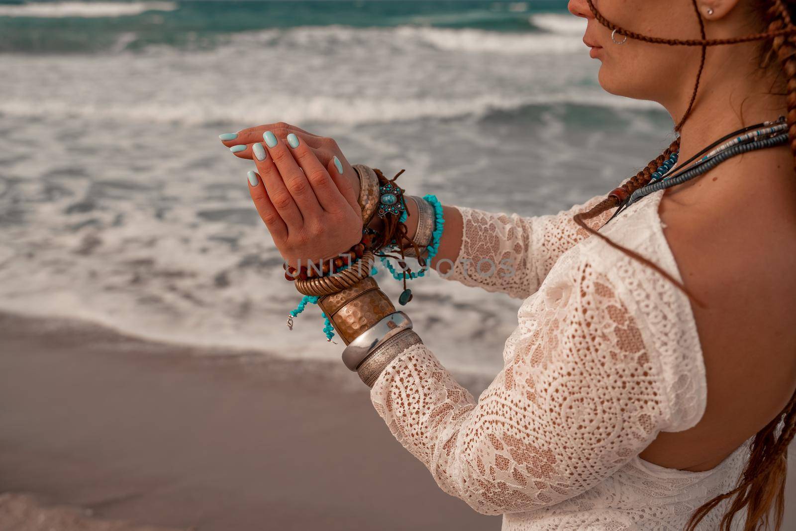 Model in boho style in a white long dress and silver jewelry on the beach. Her hair is braided, and there are many bracelets on her arms