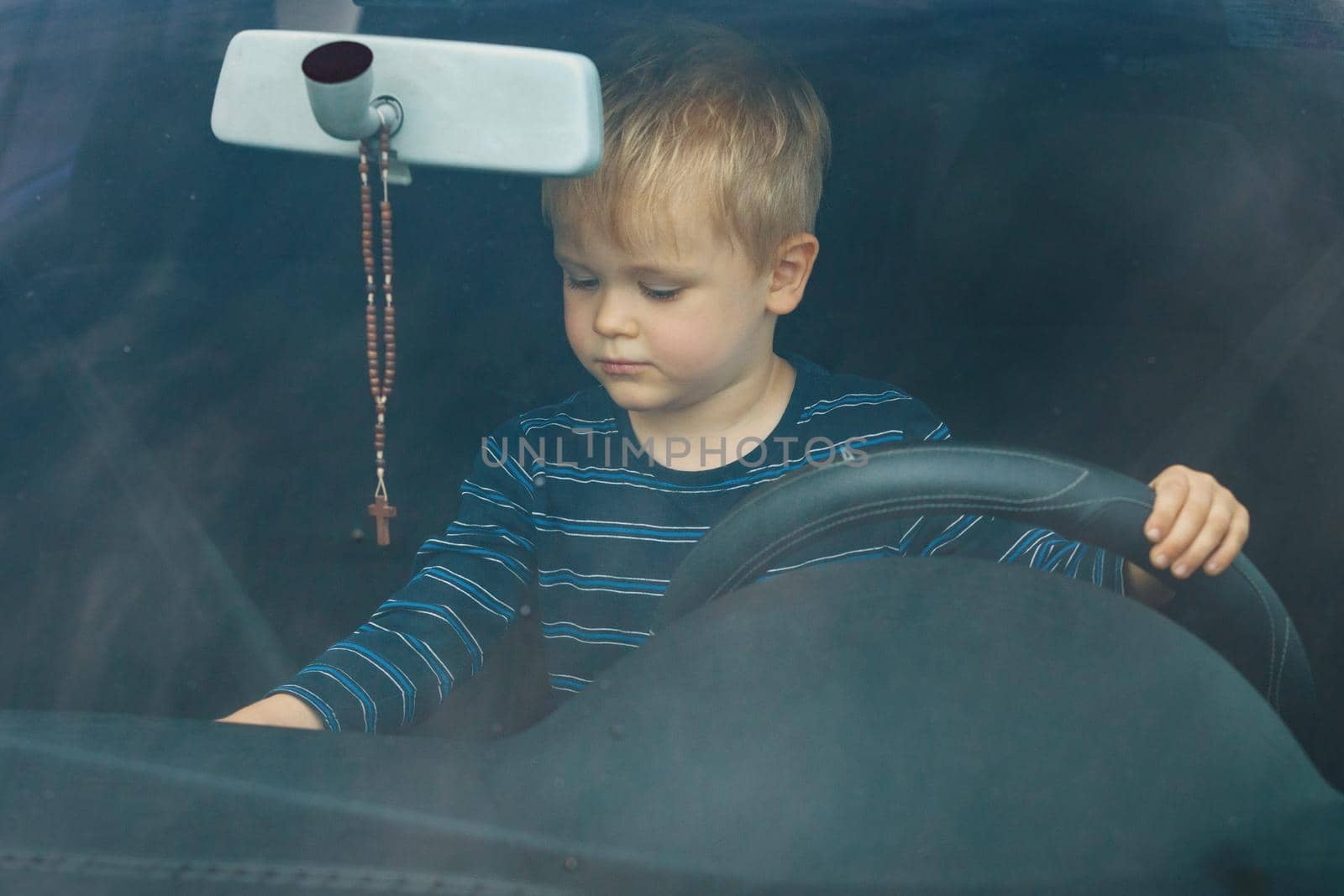 Cute little boy driving fathers car. Portrait of a child sitting in a car behind the wheel from the front. The boy plays and imagines he is driving, he controls the radio with the other hand.