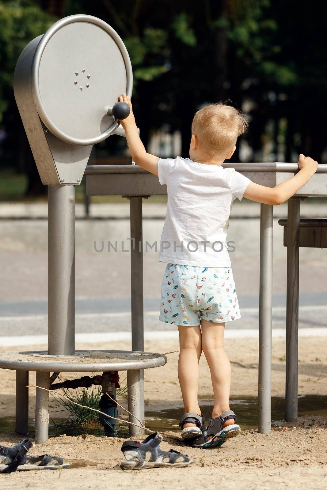 Barefoot little boy wearing shorts and t-shirt playing with water pump faucet in public park.