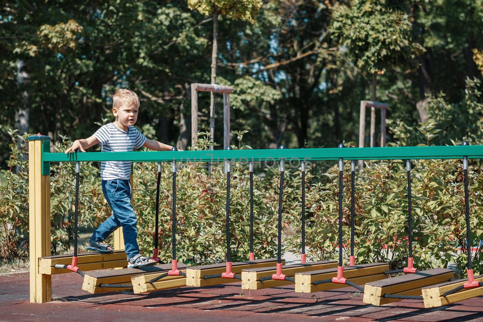 A brave little boy goes across the bridge of the monkeys in a city playground, against a green foliage background.