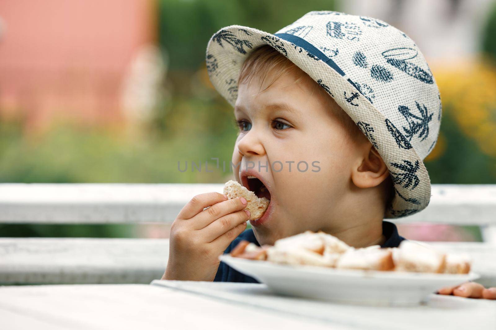Close up image of Caucasian baby boy with blonde hair and hat opening mouth going to bite crispy bread, having joyful facial expression. by Lincikas