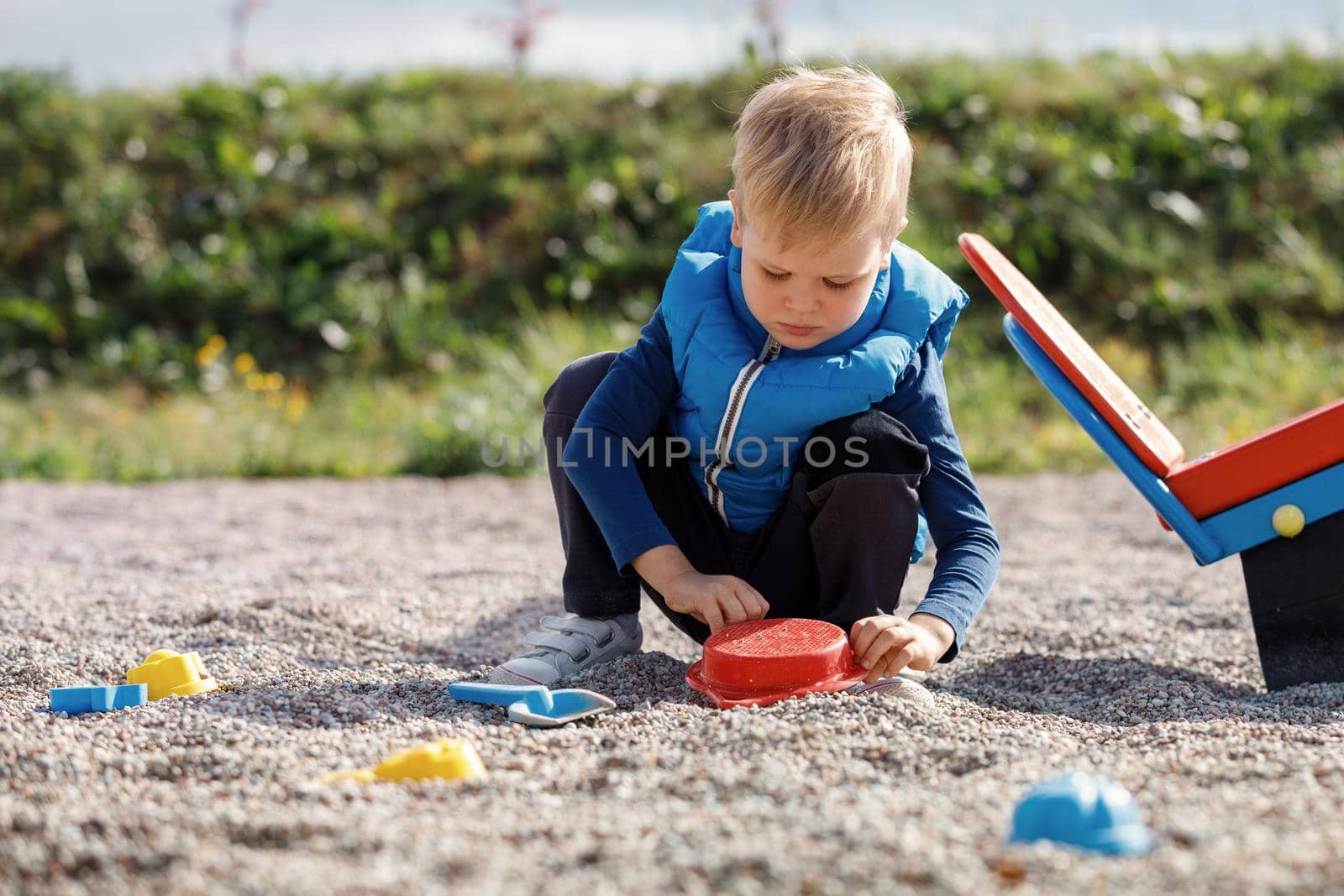 A cute blond child is playing with sand toys near the balance swing in the yard. Safe surface with of small pebbles in playground. by Lincikas