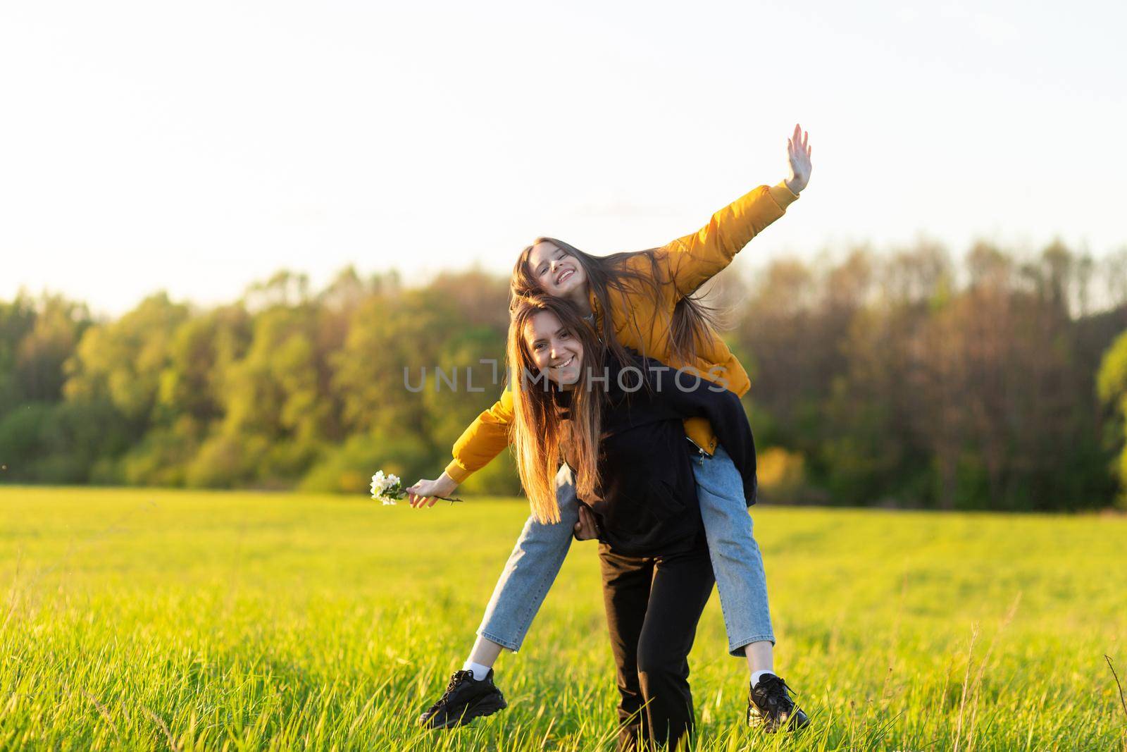 Playful mother giving daughter piggy back ride at green field. Both laughing and look happy. Spring in forest background. Closeup.