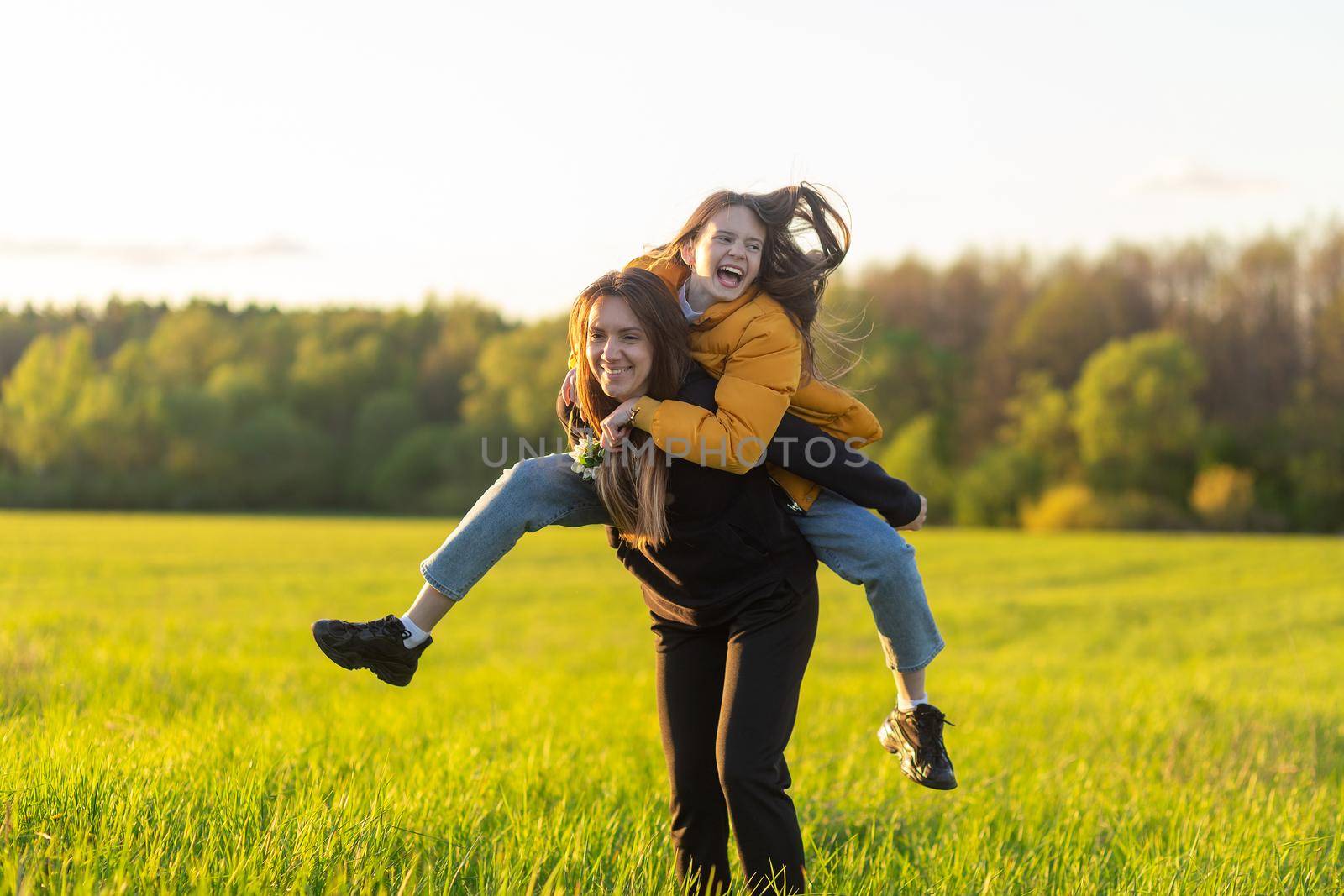 Playful mother giving daughter piggy back ride at green field. Both laughing and look happy. Spring in forest background. Closeup.