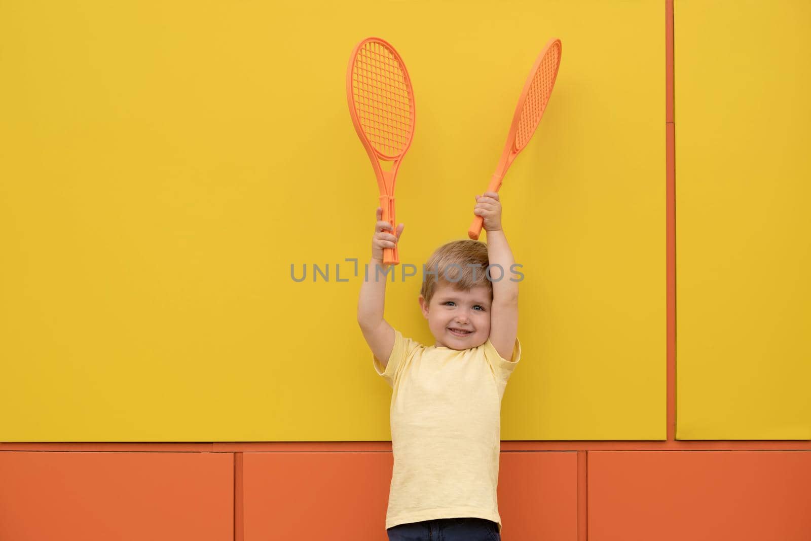 A boy with tennis rackets against yellow wall