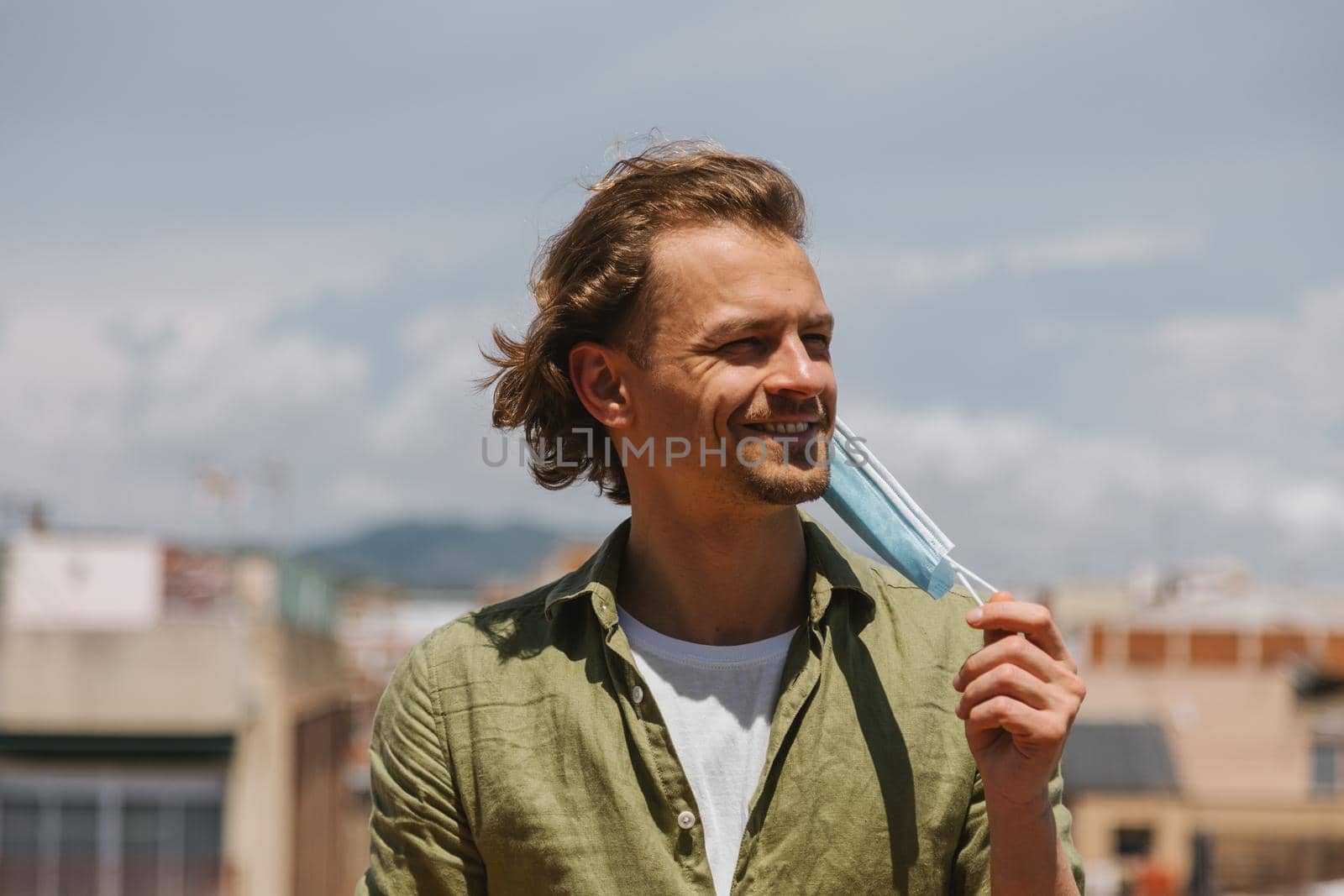 End of coronavirus pandemic. European man is happy to take off medical mask on sunny day with city and sky background. by apavlin