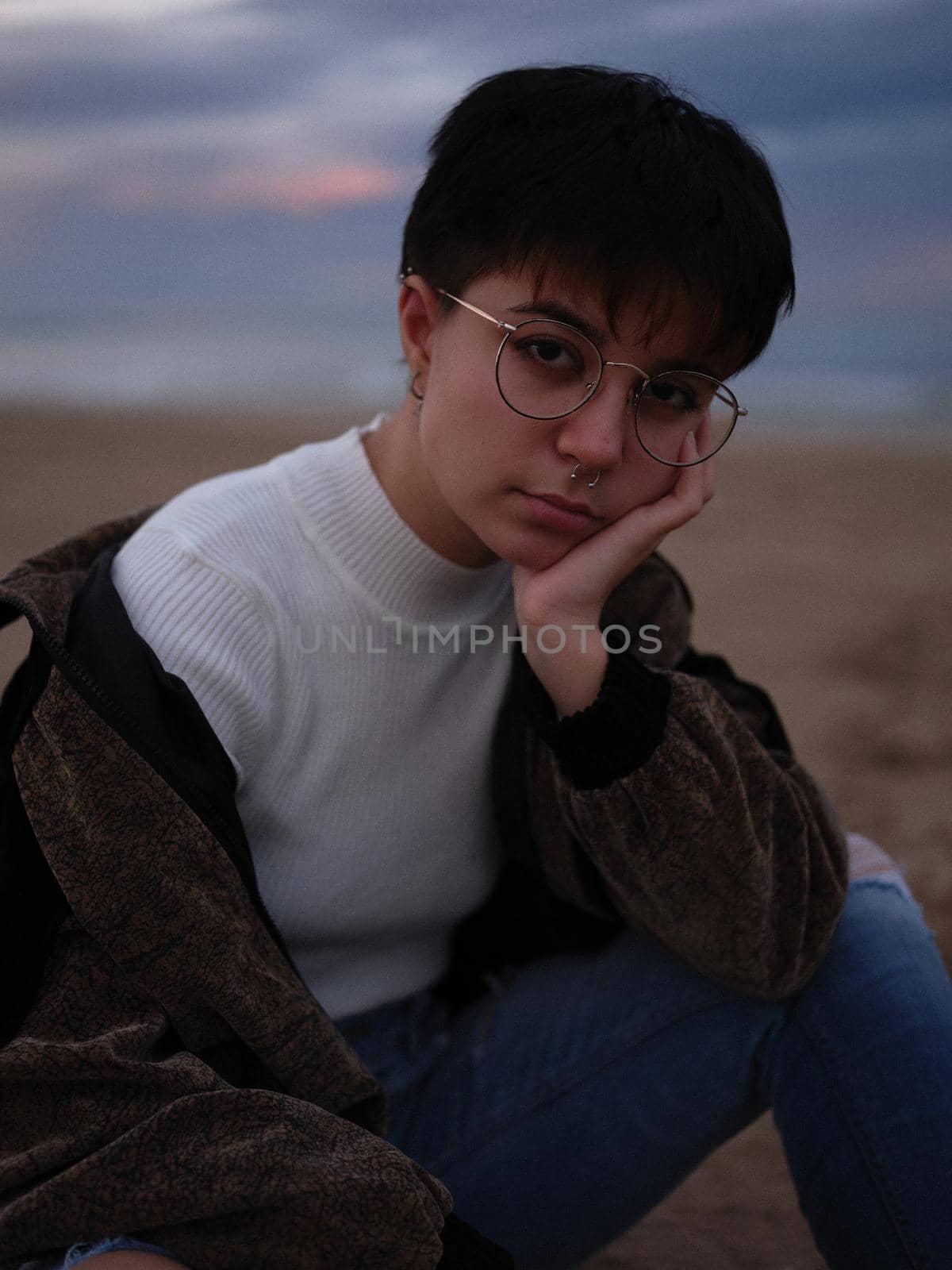 tired transsexual woman sitting on the beach holding her head with her arm, vertical portrait on a blurred background