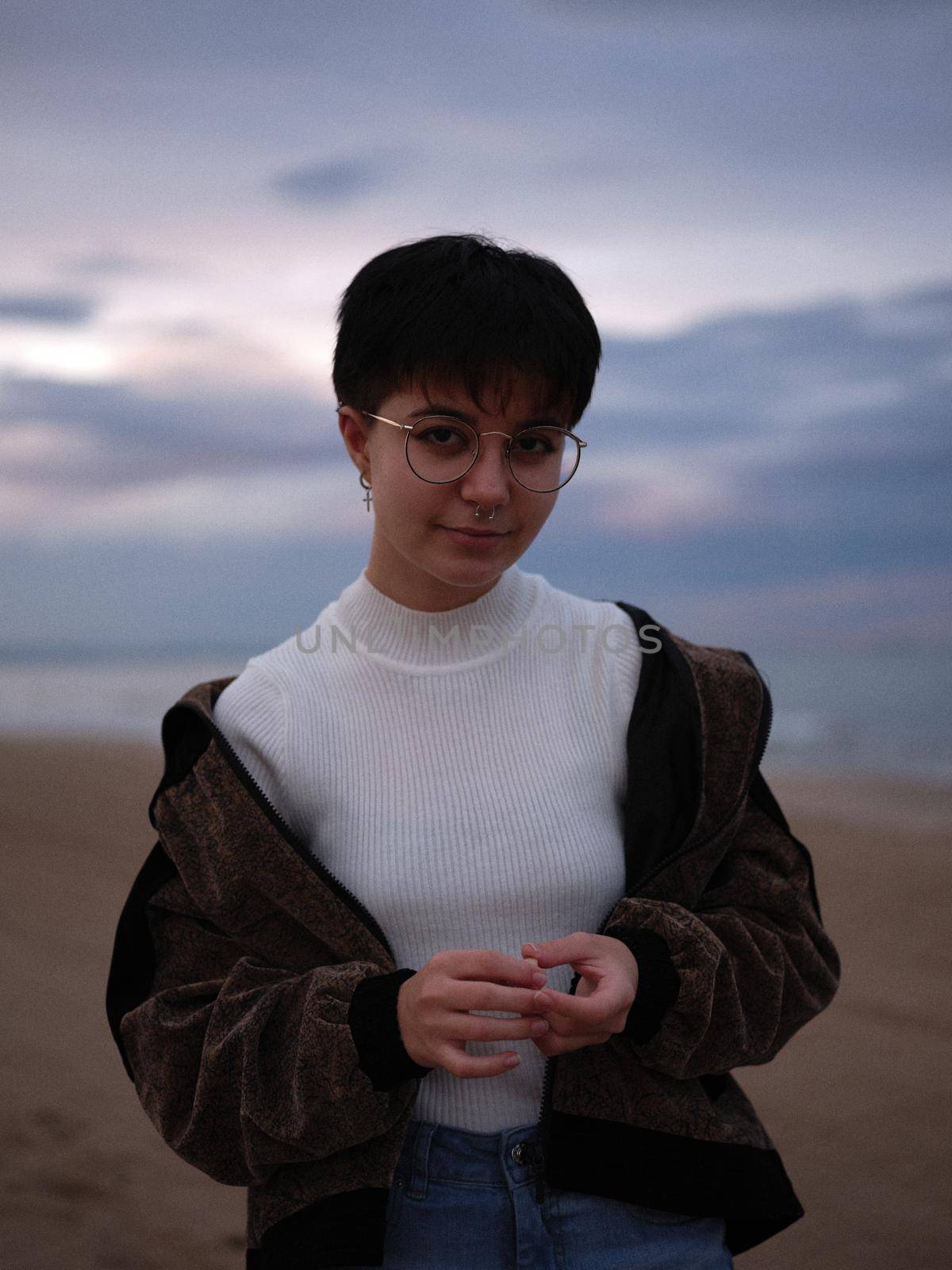 smiling non-binary girl standing looking at the camera with the beach behind her, background out of focus