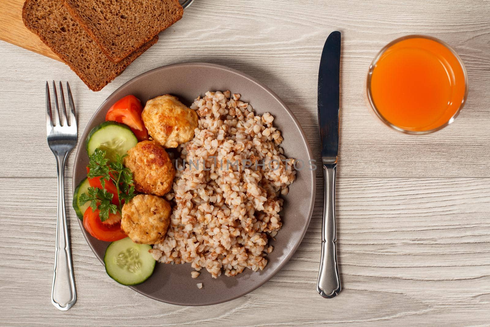 Plate with buckwheat, fried meat cutlets, pieces of fresh cucumbers, tomatoes and fresh parsley with knife and fork, bread on wooden cutting board and glass of orange juice. Top view