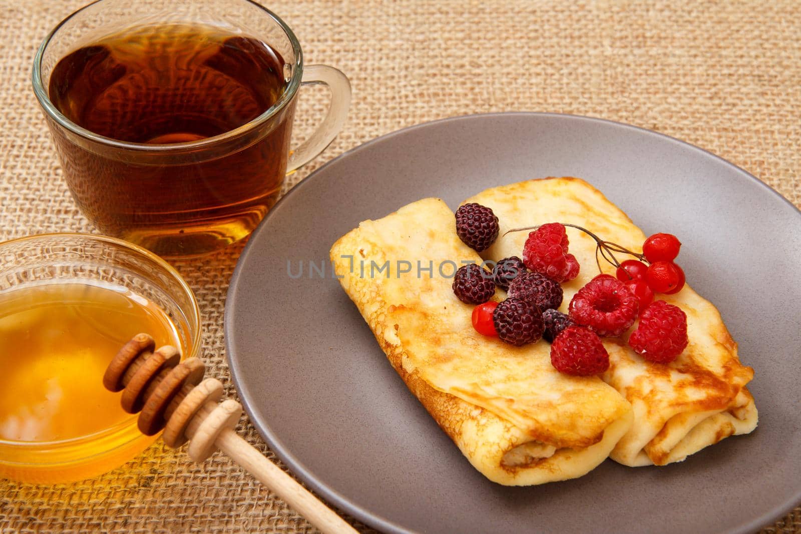 Homemade pancakes filled with cottage cheese and topped with frozen raspberries and blackberries on plate, cup of tea and honey in glass bowl with wooden spoon on sackcloth