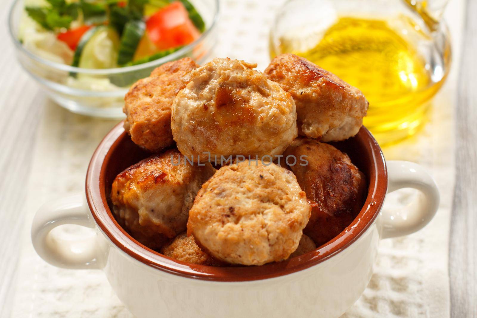Fried meat cutlets in ceramic soup bowl, bottle with sunflower oil and salad of fresh cucumbers and tomatoes in glass bowl on white kitchen towel