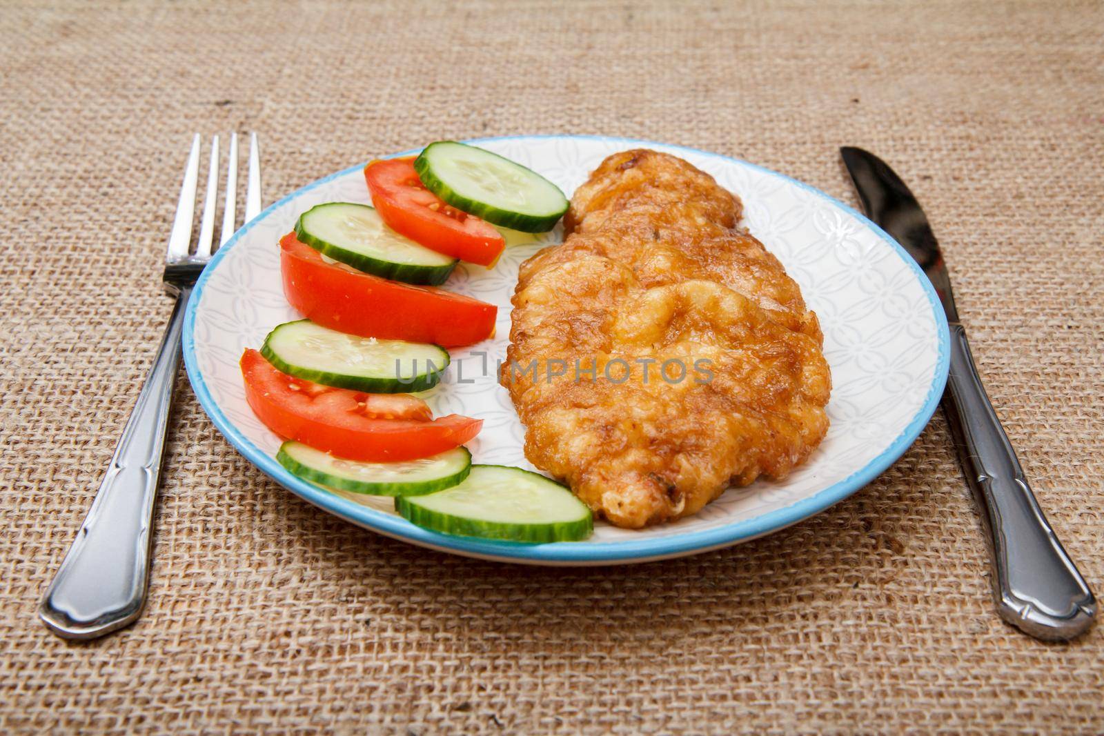 Plate with fried meat chop in egg batter and sliced tomato and cucumber with knife and fork on sackcloth