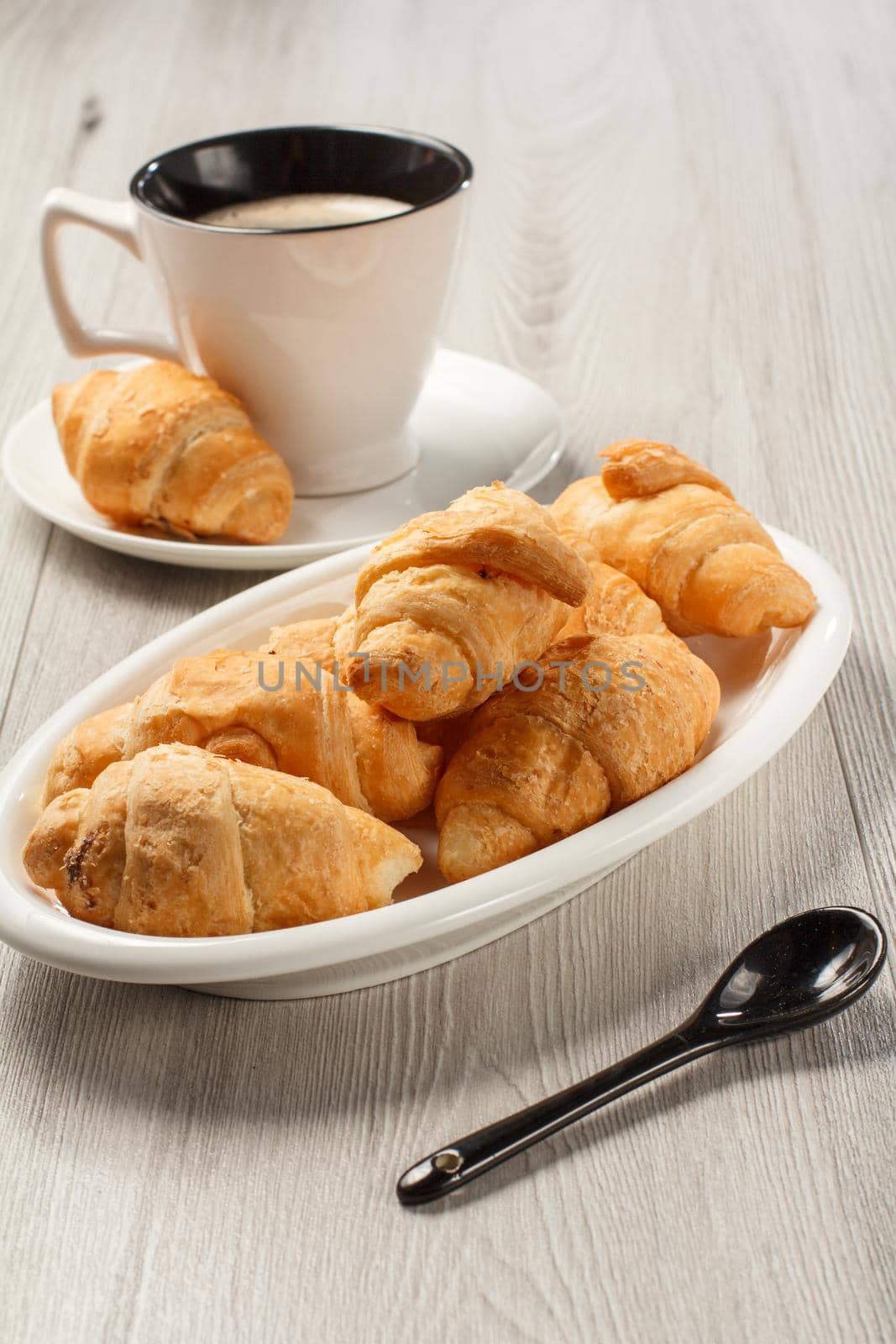 Fresh croissants on white porcelain dish, cup of black coffee with saucer on background