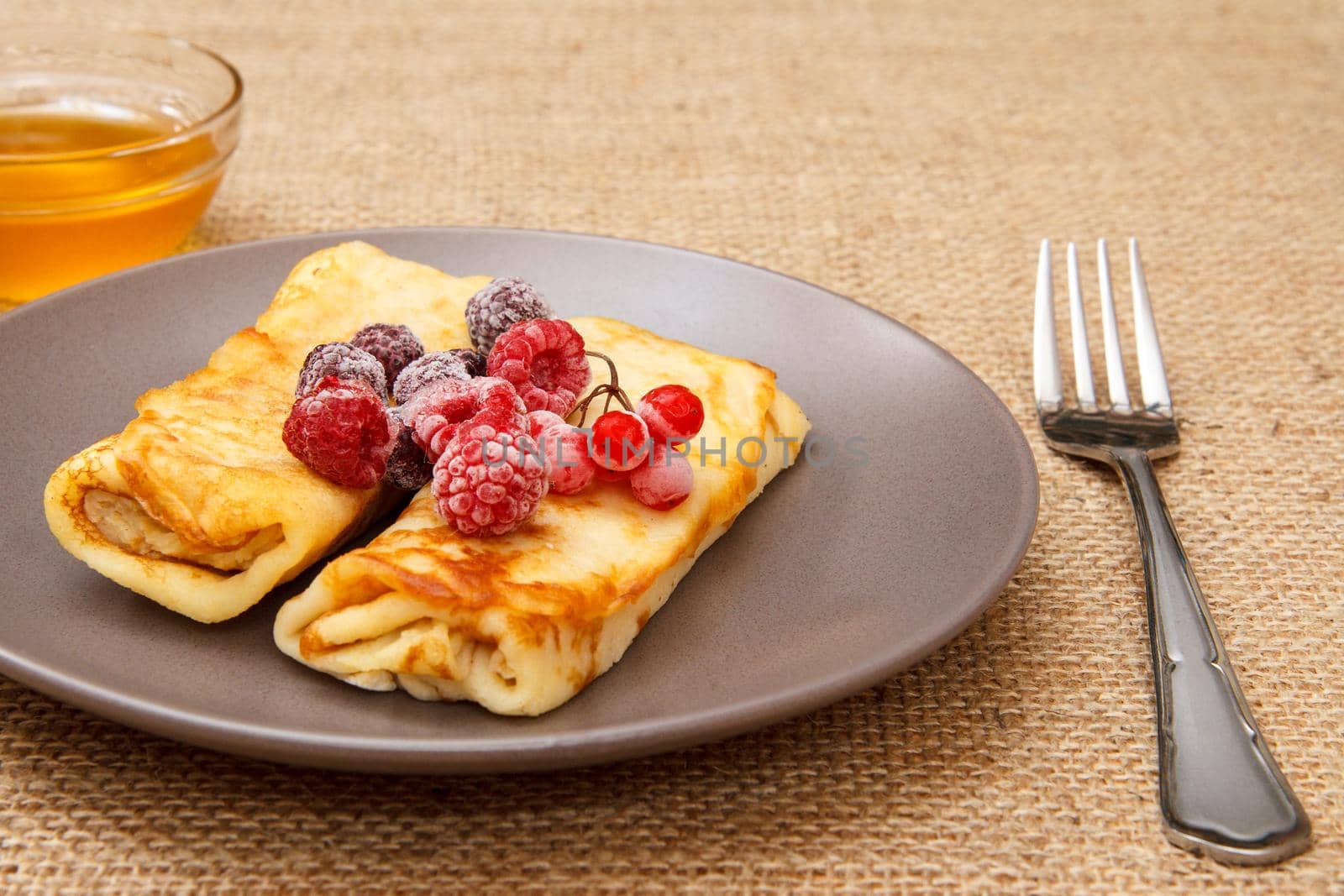 Homemade pancakes filled with cottage cheese and topped with frozen raspberries and blackberries on plate, honey in glass bowl and fork on sackcloth background