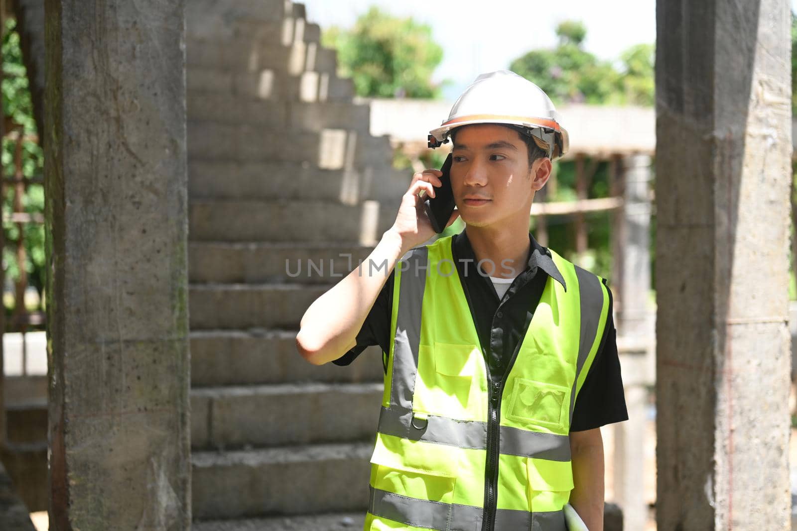 Young engineer builder in helmet talking on mobile phone while standing at construction site.