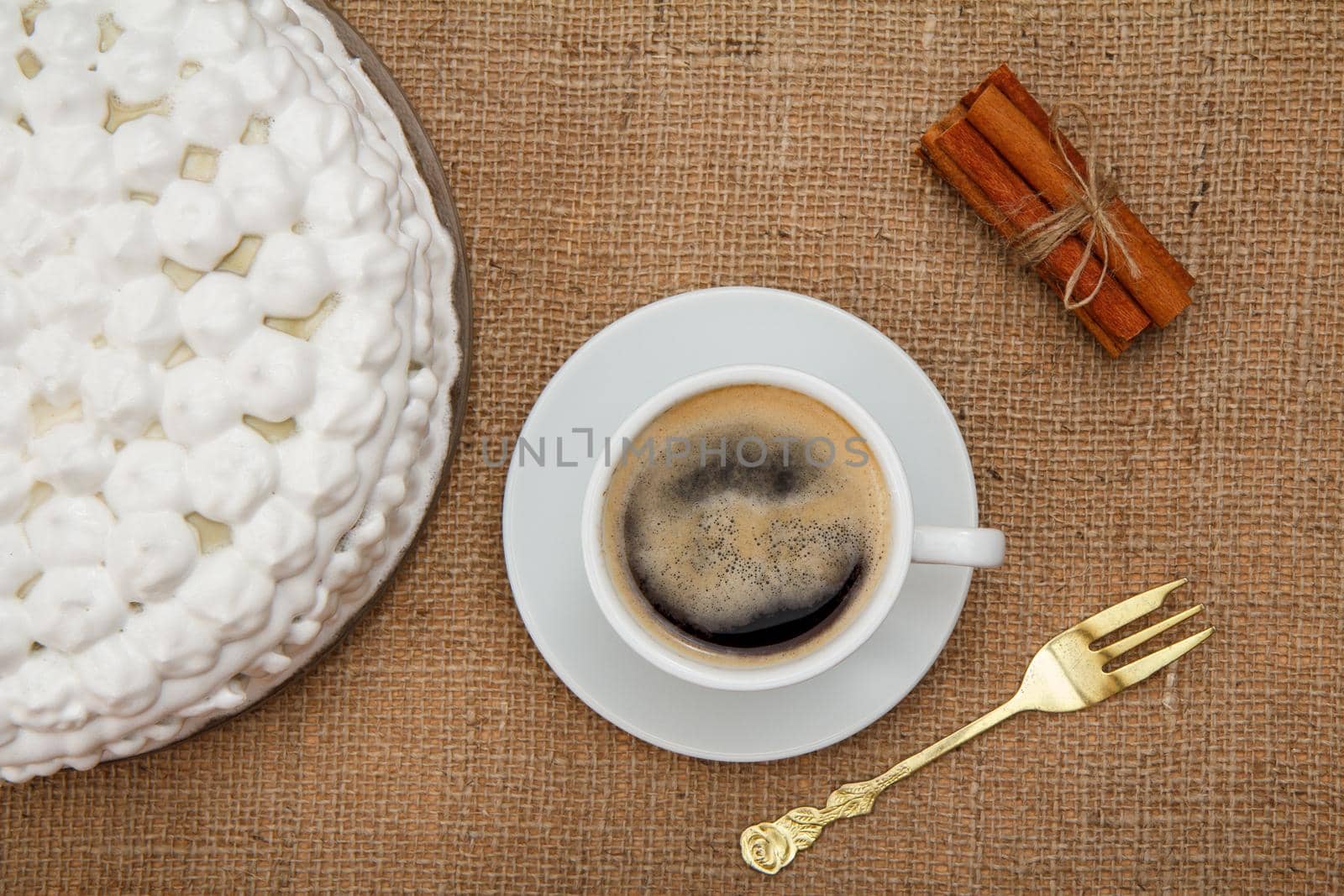 Biscuit cake decorated with whipped cream, cup of coffee, fork and cinnamon on table with sackcloth. Top view