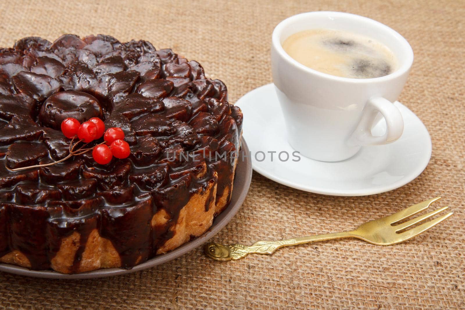Chocolate cake decorated with bunch of viburnum, fork and cup of coffee beside it on table with sackcloth