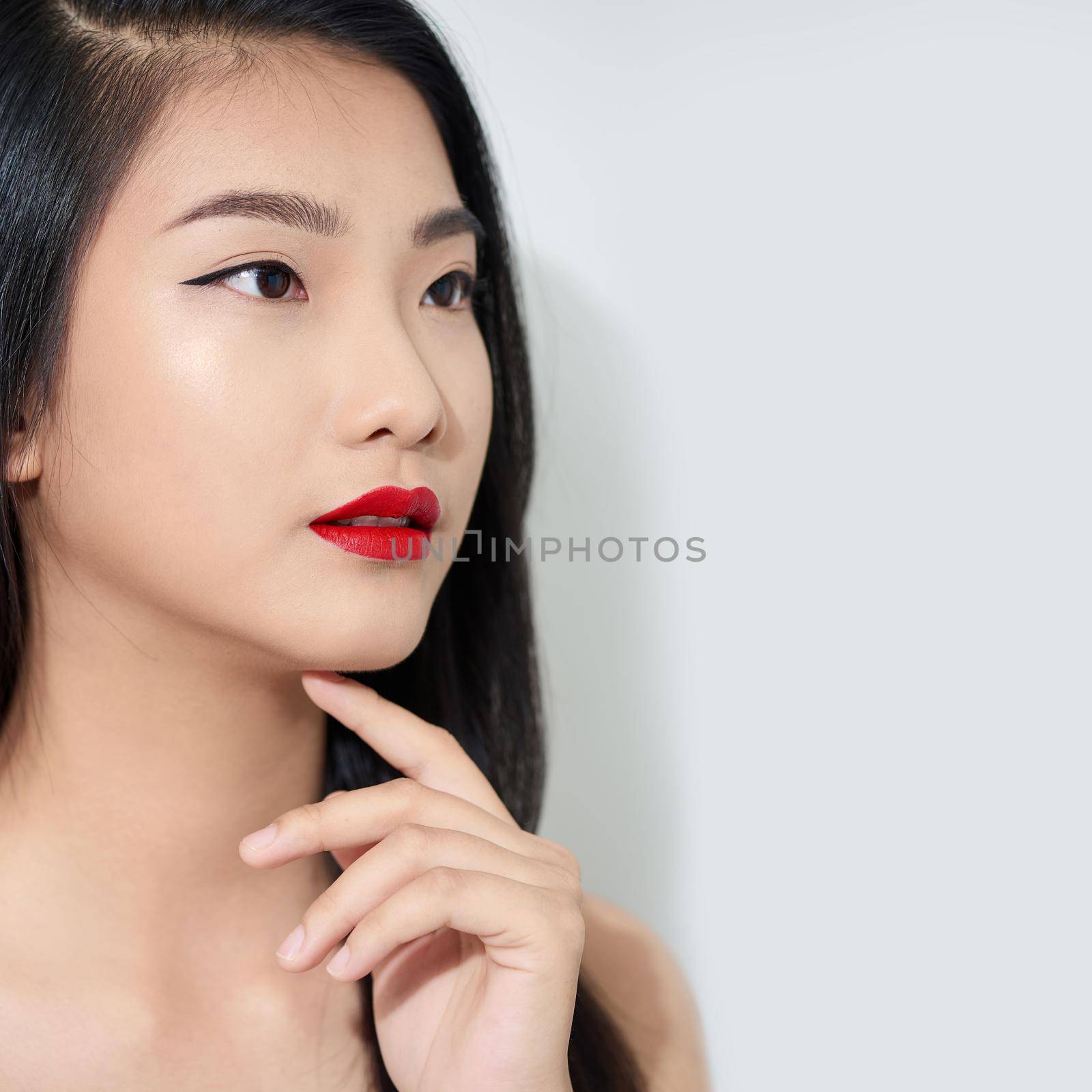 Beautiful stylish young woman is touching her chin, looking away and smiling, on white background