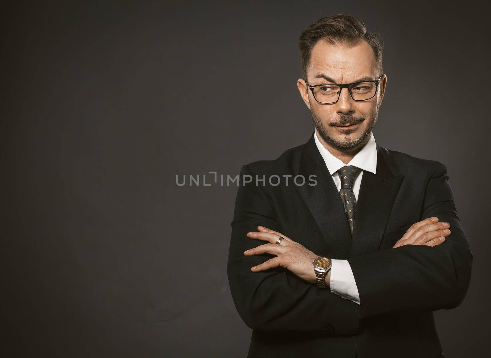 Confident businessman cunningly looks away with his arms crossed. Caucasian middle-aged man standing against a gray wall with copyspace or space for text on the left. Toned image.