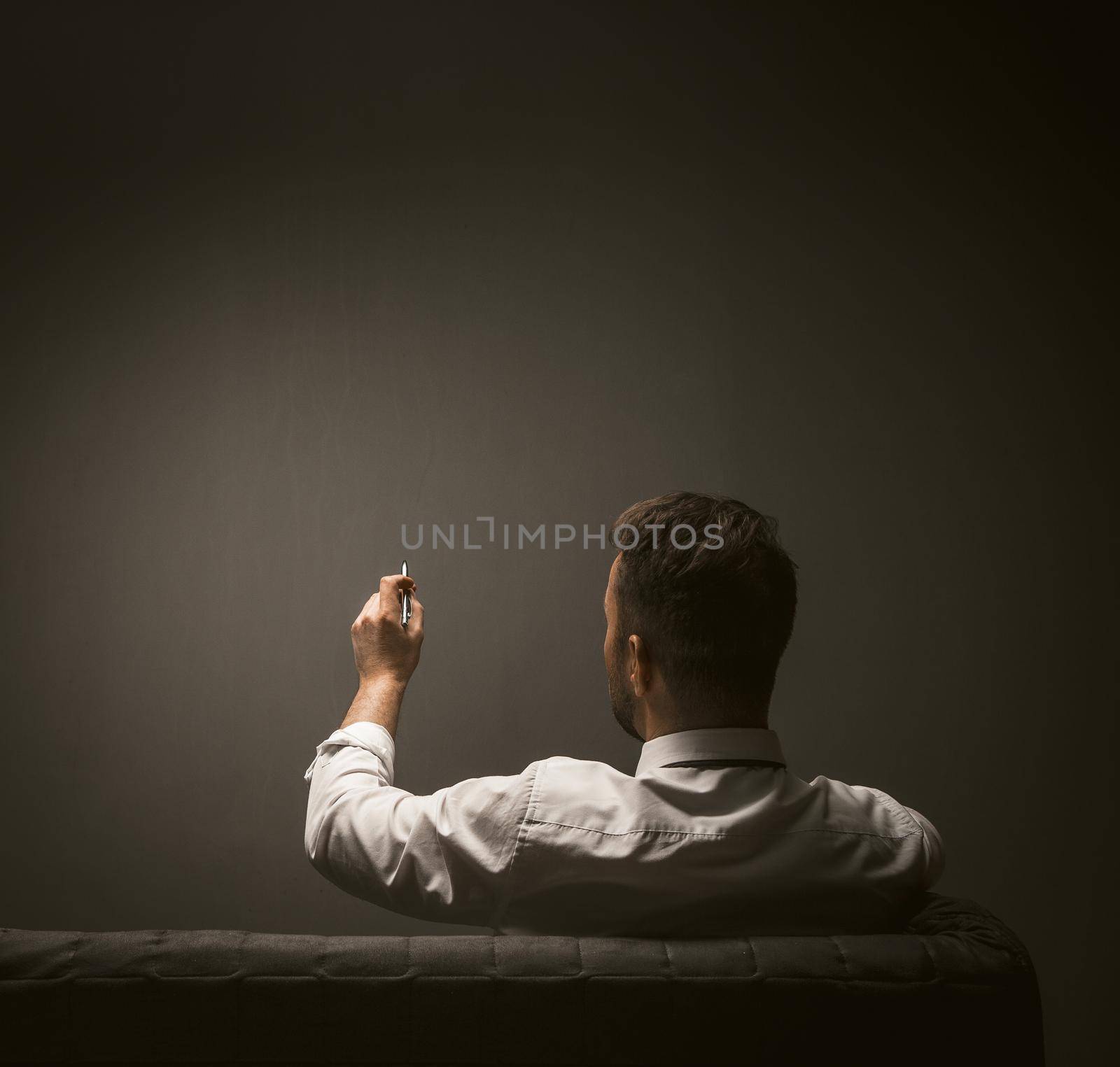 Businessman writing with pen on dark gray wall. View from the back of man in a white shirt makes notes while sitting on couch. Blank or template for creativity. Toned image.