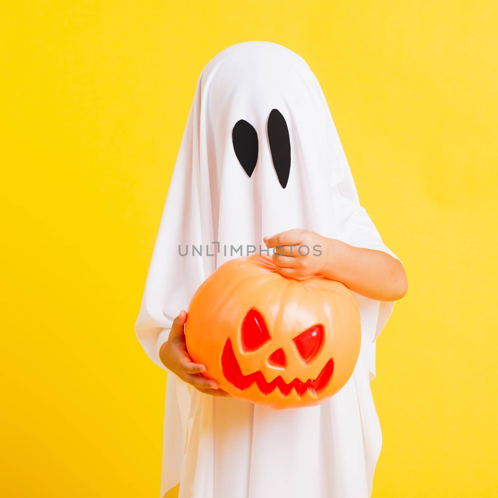 Funny Halloween Kid Concept, little cute child with white dressed costume halloween ghost scary he holding orange pumpkin ghost on hand, studio shot yellow on white background