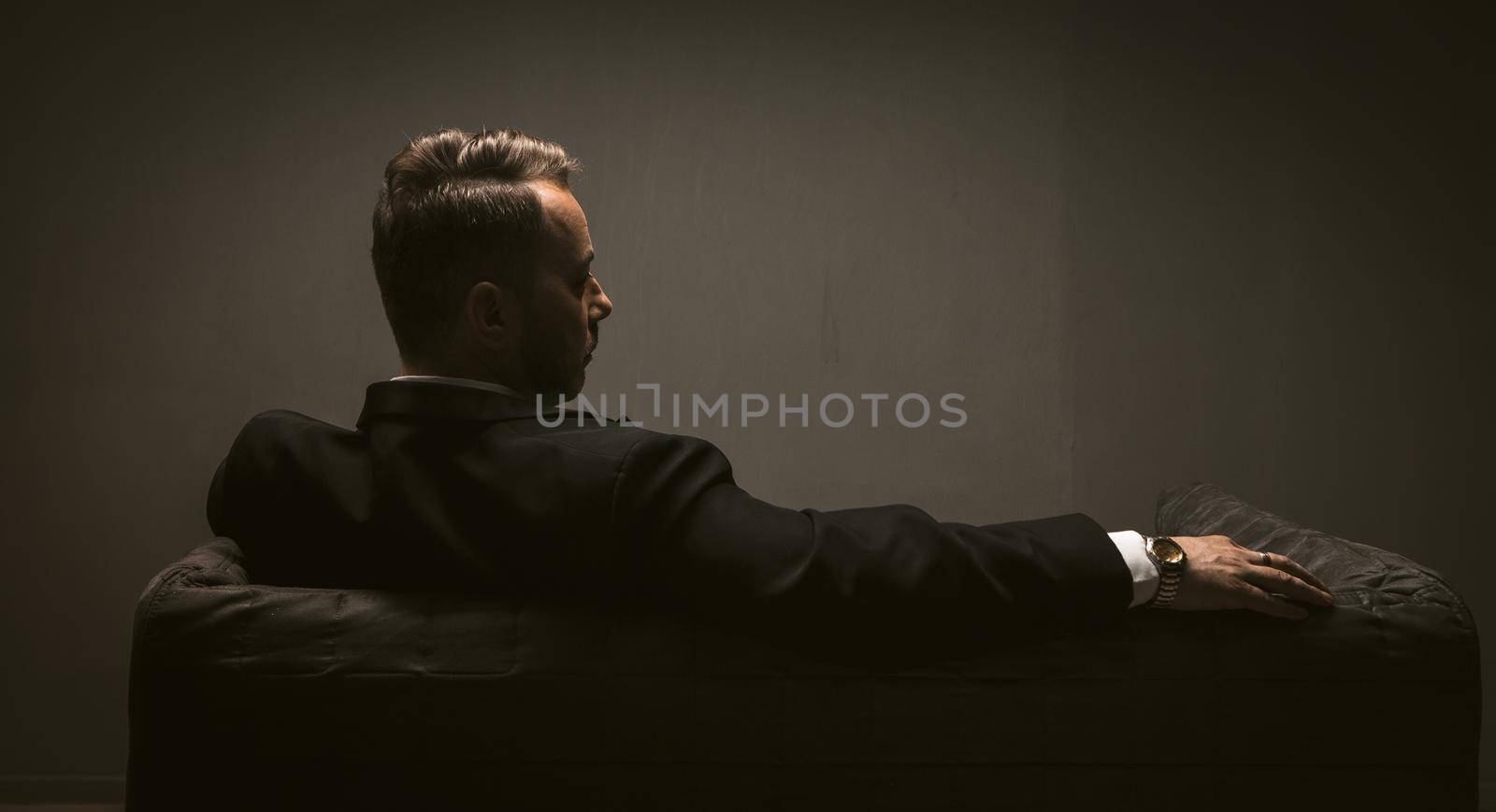 Thoughtful businessman sits at sofa in dark room. Rear view of pensive elegant man is alone with his thoughts. Lonelinessconcept. Leadership concept. Toned image by LipikStockMedia