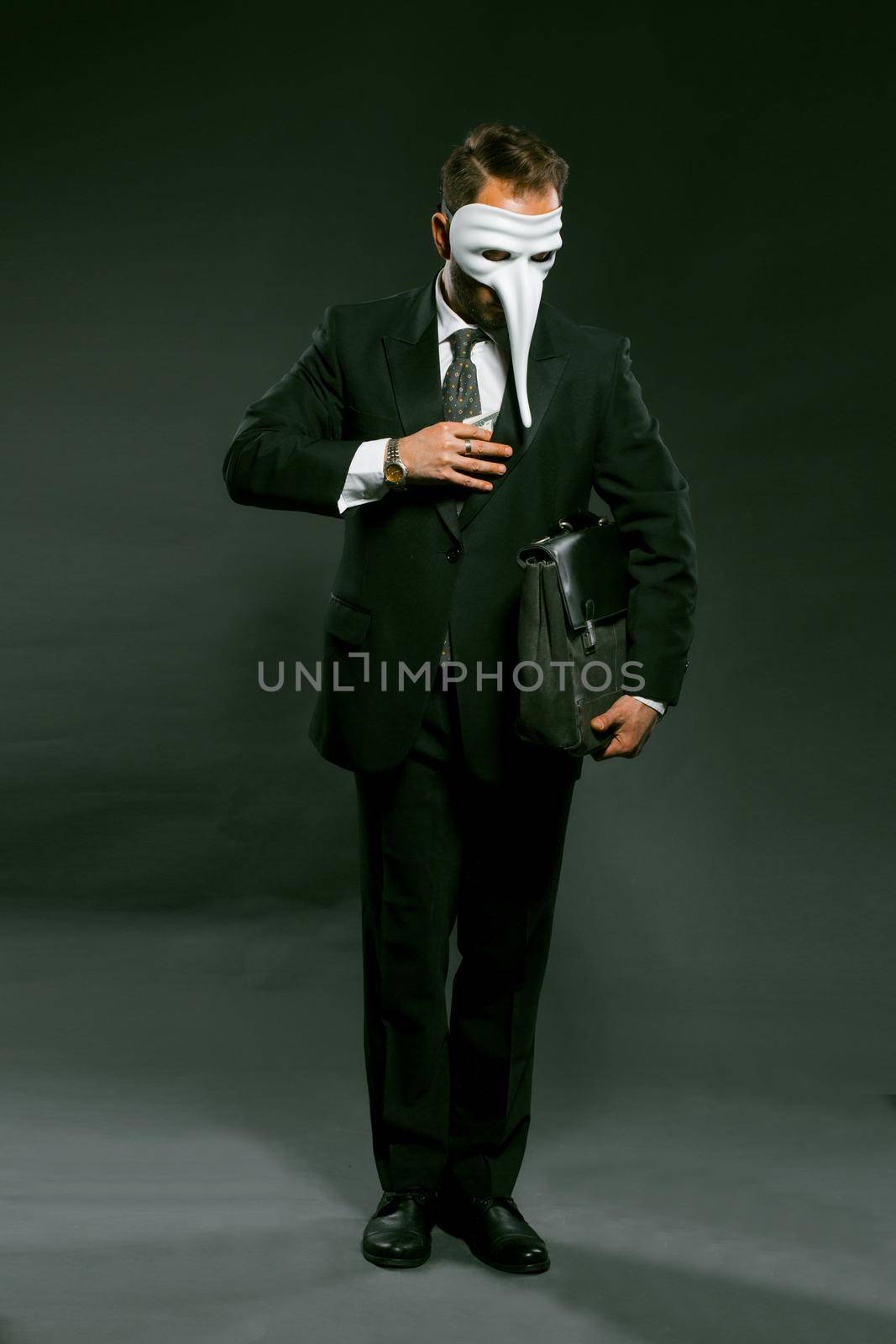 Masked businessman puts large wad of money in his breast pocket holding briefcase under his arm. Man in fancy white masks full-length stands on gray background. Corruption concept. Toned image by LipikStockMedia